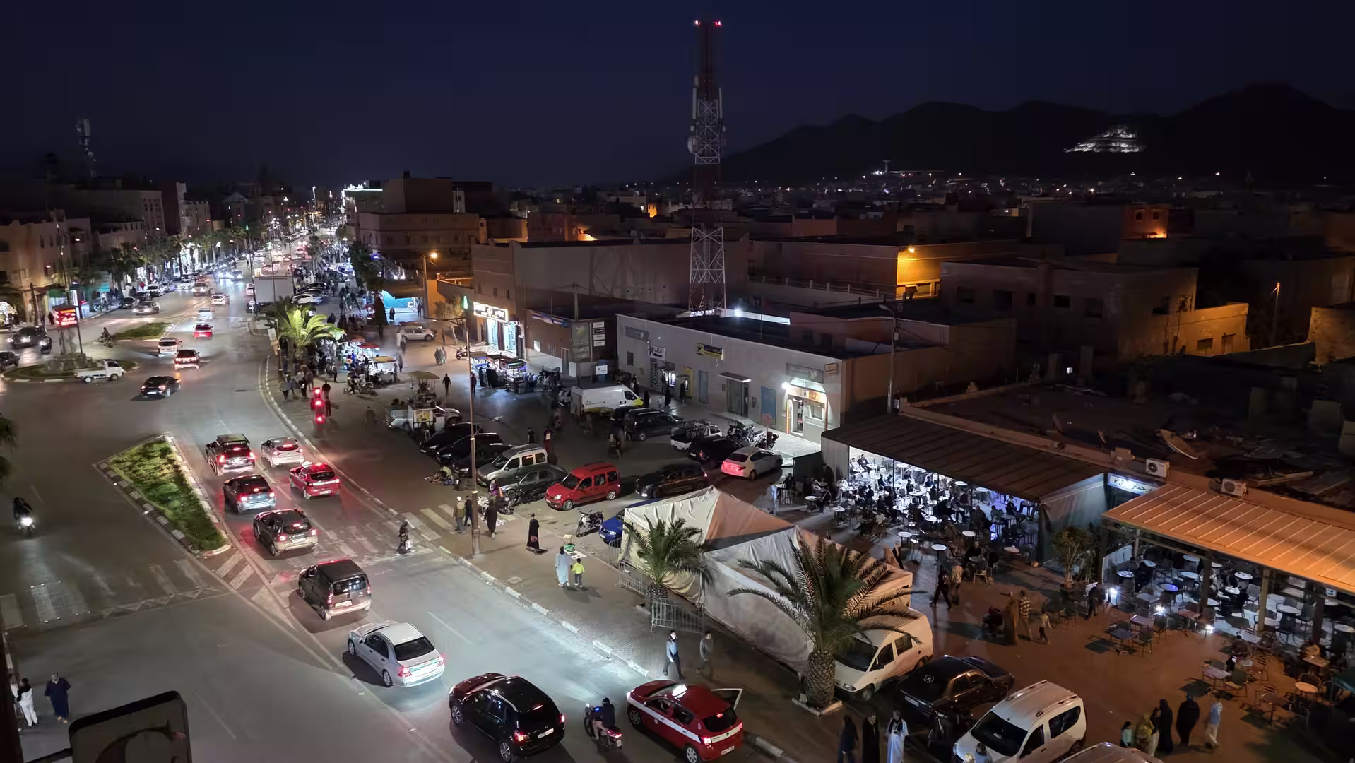 Elevated nighttime view across central Guelmim, Morocco, showing busy streets, restaurants, parked cars, illuminated storefronts and the dark mountain skyline beyond.