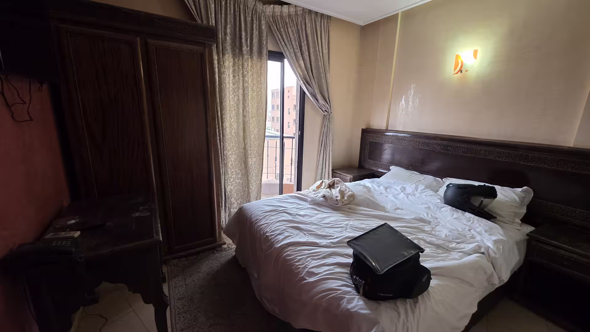 Interior of a traditional-style hotel room in Guelmim, Morocco, with carved wooden furniture, an unmade double bed and a balcony door with curtains.