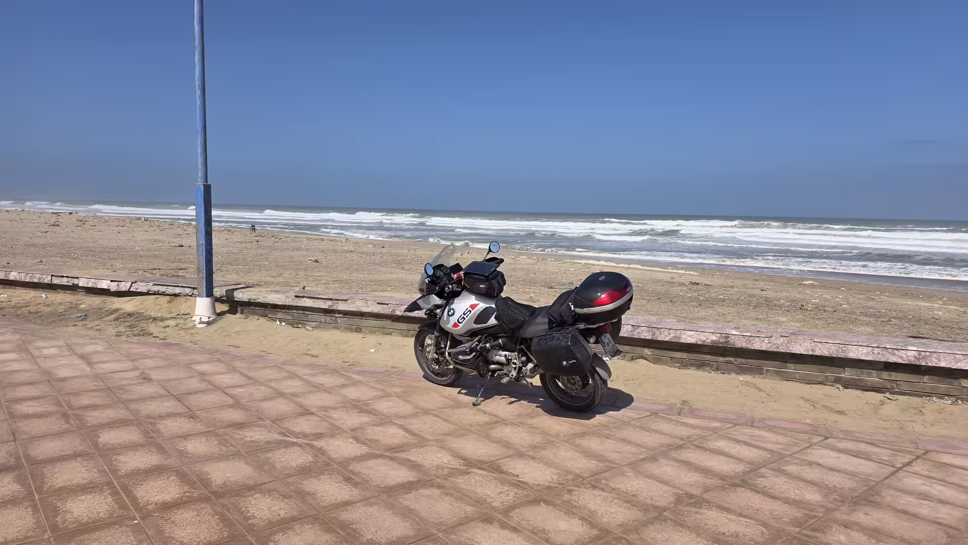 Touring motorcycle parked on a seaside promenade beside a sandy Atlantic beach with rolling waves under a clear blue sky in southern Morocco.