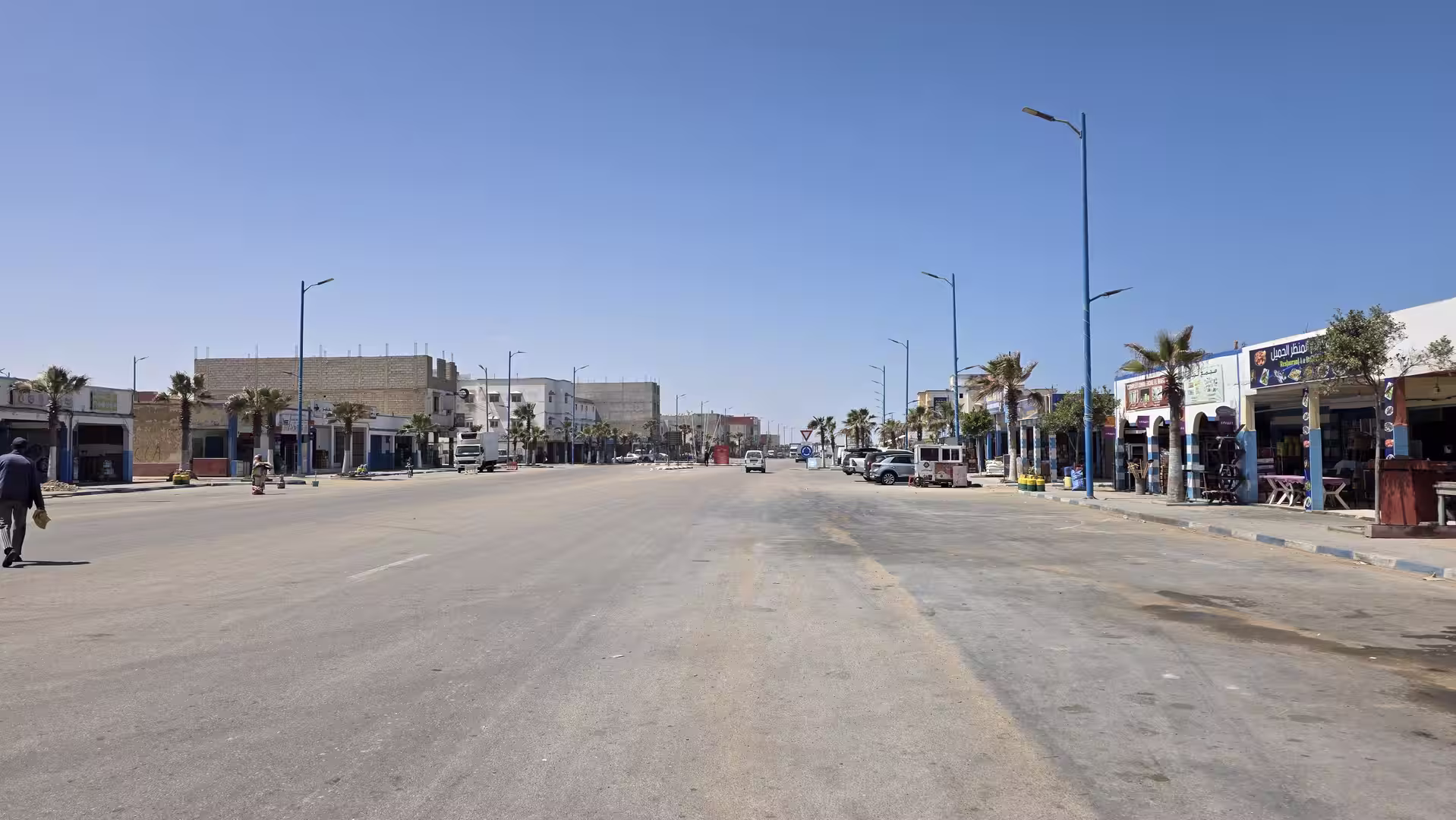 Wide boulevard in a coastal town in southern Morocco with palm-lined sidewalks, low-rise buildings, shops and clear blue skies.