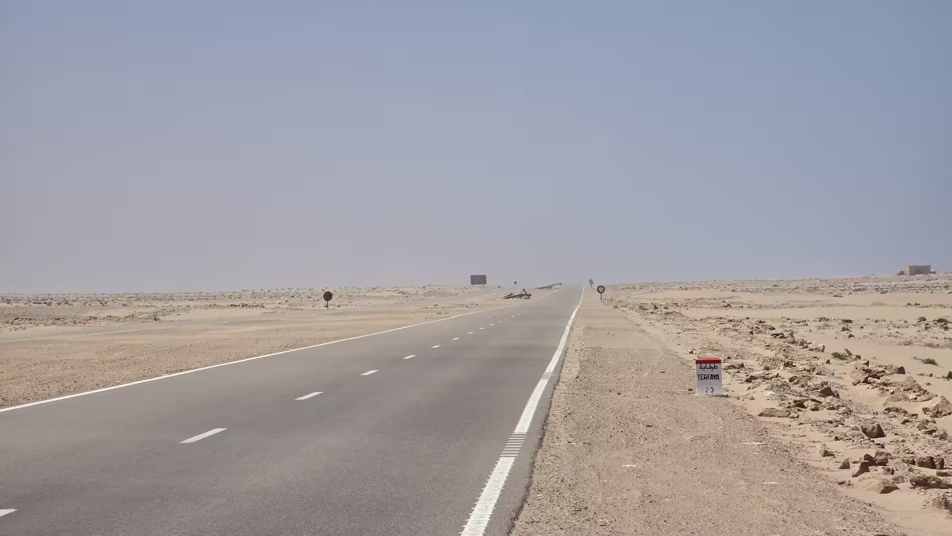 Straight asphalt road crossing a flat desert landscape in southern Morocco with sparse rocky ground and a roadside marker indicating Tarfaya 23 kilometres ahead.