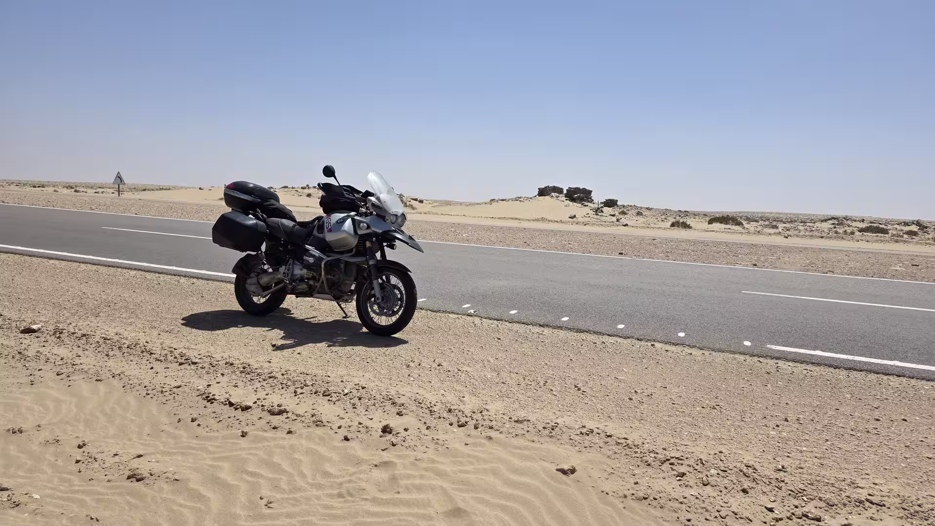 Adventure motorcycle parked beside a paved road in a sandy desert landscape near Tarfaya in southern Morocco under a clear blue sky.