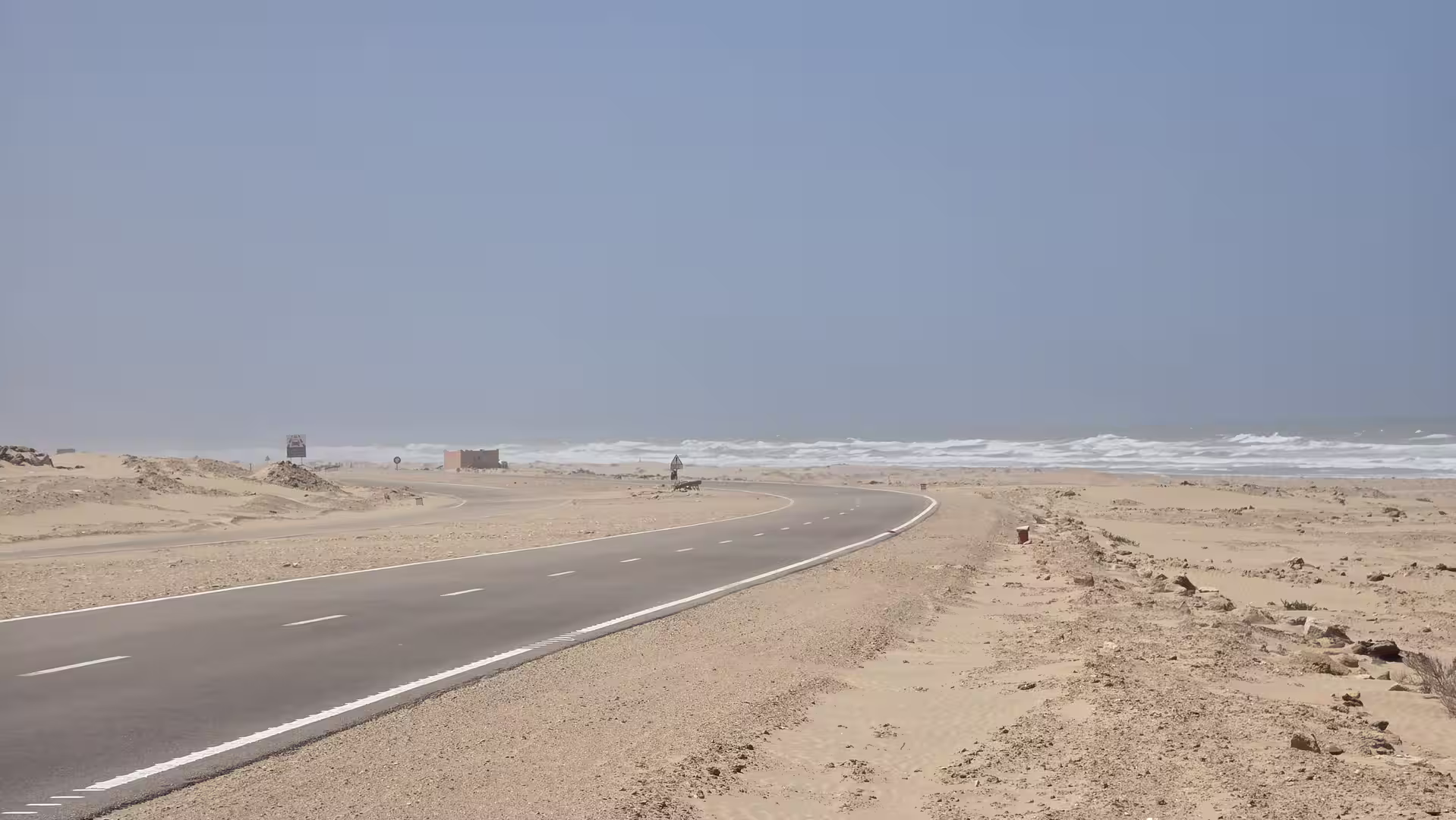 Curving coastal highway running through sandy desert terrain beside the Atlantic Ocean near Tarfaya in southern Morocco, with waves visible in the background.
