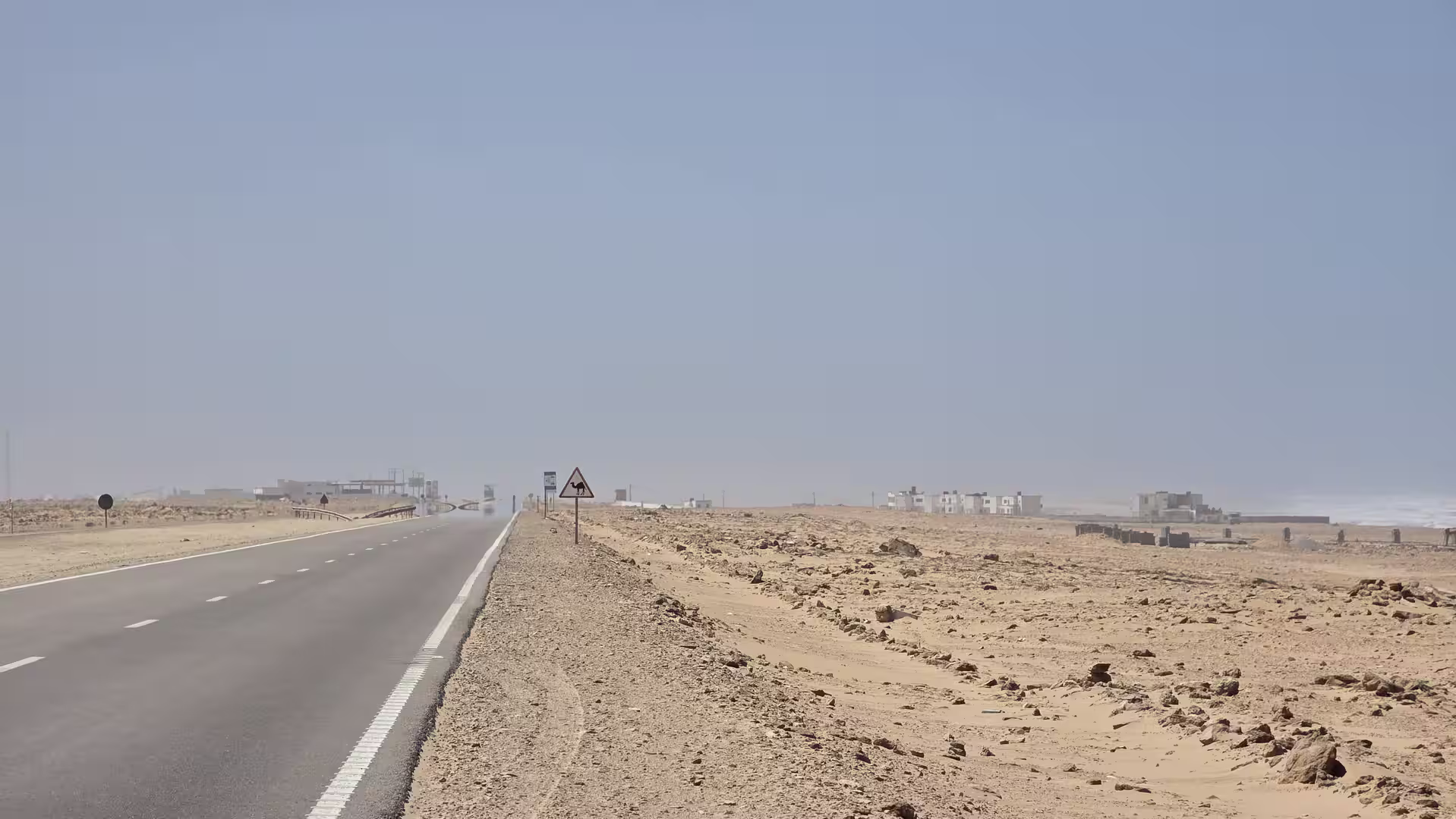 Desert highway running along the Atlantic coast in southern Morocco with a camel crossing warning sign and scattered buildings in the distance.