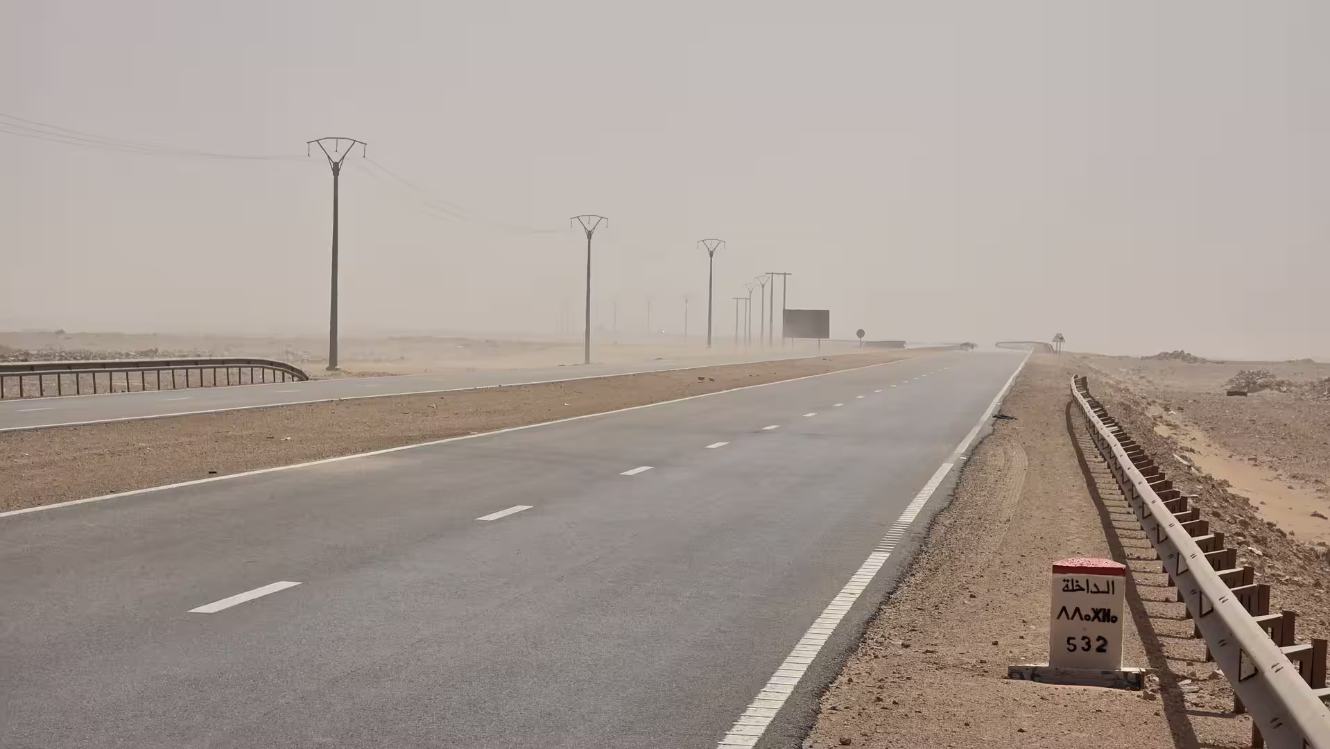 Desert highway under hazy sand-filled conditions in southern Morocco with power lines, roadside barriers and a kilometre marker indicating Dakhla 532 kilometres away.