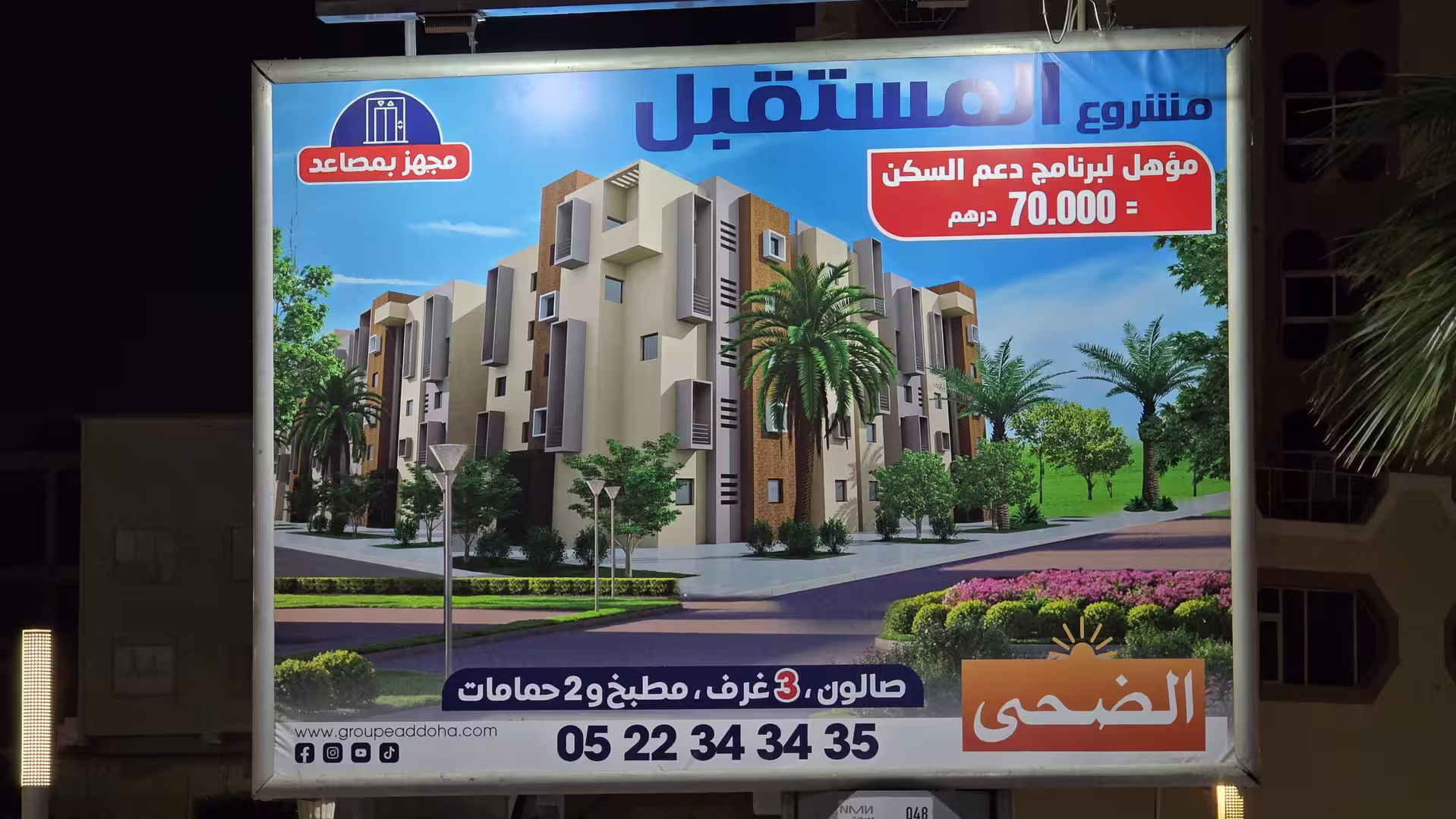 Illuminated billboard at night in Laâyoune advertising a residential apartment development with a rendering of apartment buildings and Arabic text.