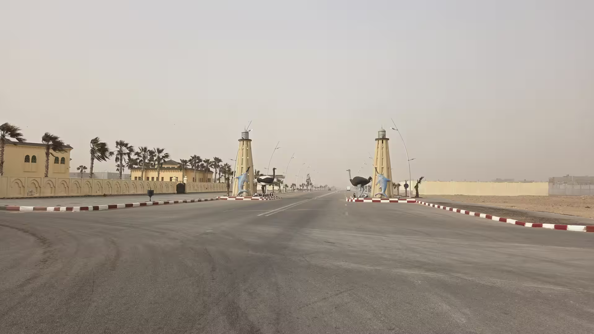 Broad entrance avenue in Boujdour, Morocco with decorative towers, palm trees and large bird sculptures under a hazy sky.