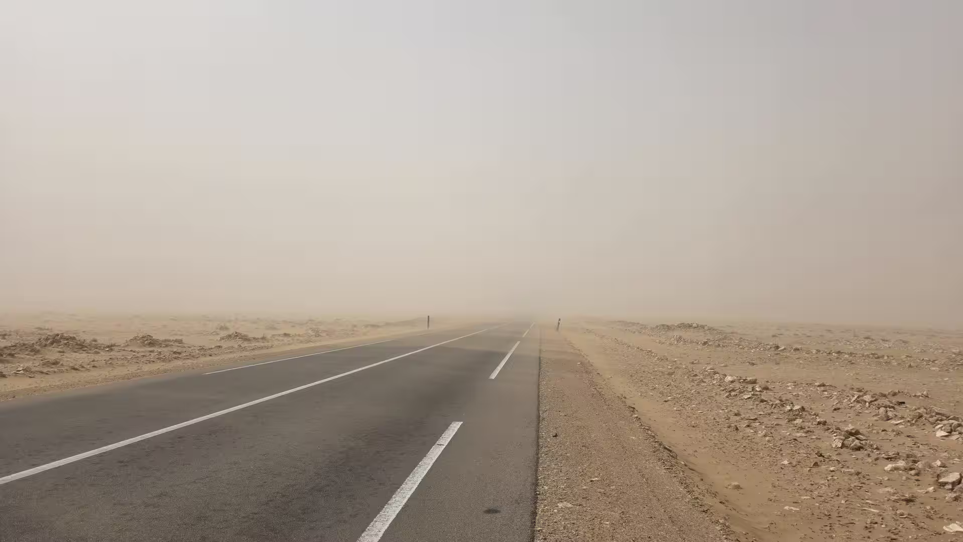 Straight asphalt road disappearing into a dusty desert horizon during a sand and haze event in southern Morocco.