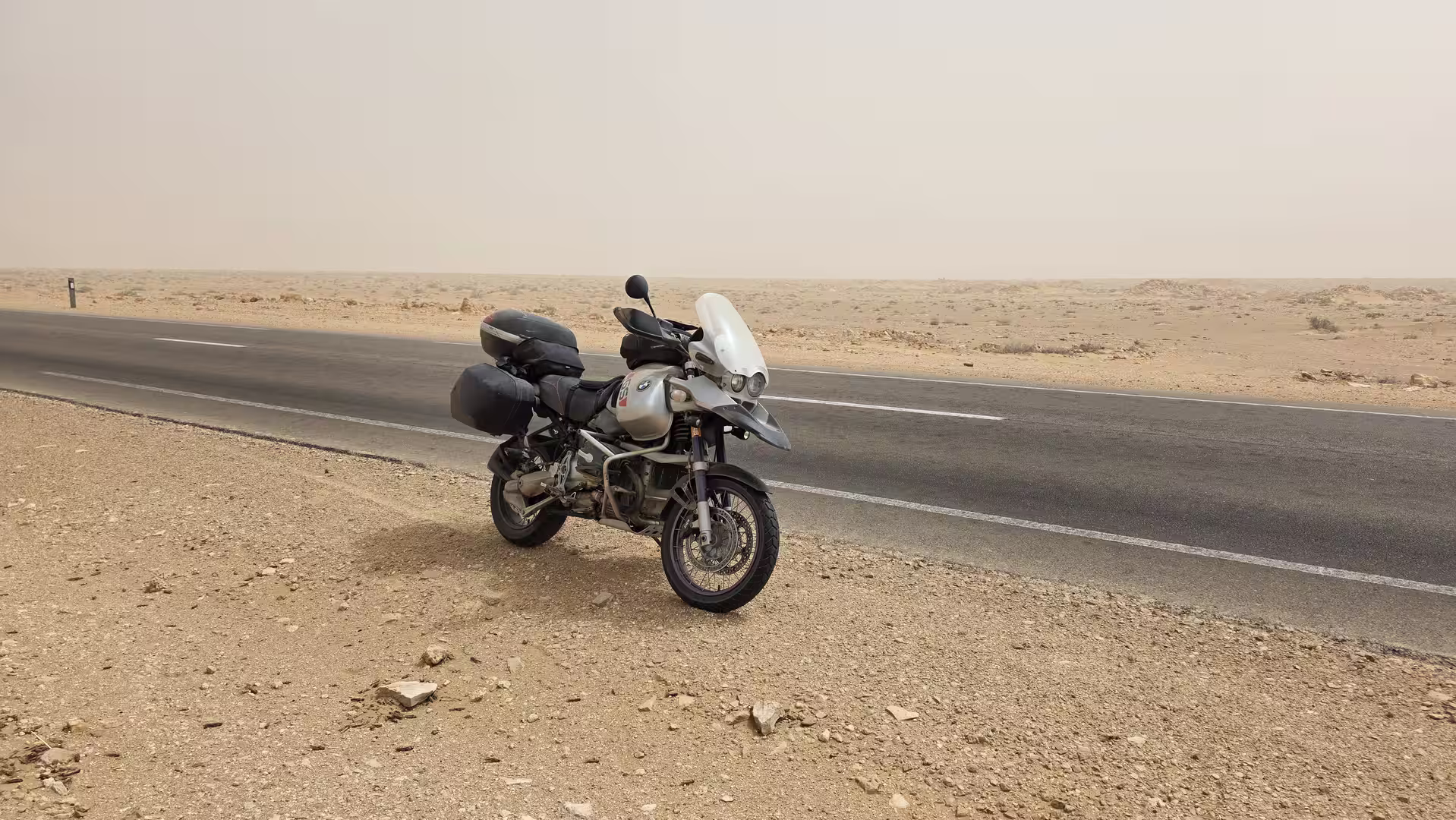Adventure motorcycle parked beside a paved road in an arid desert landscape under dusty and hazy conditions in southern Morocco.