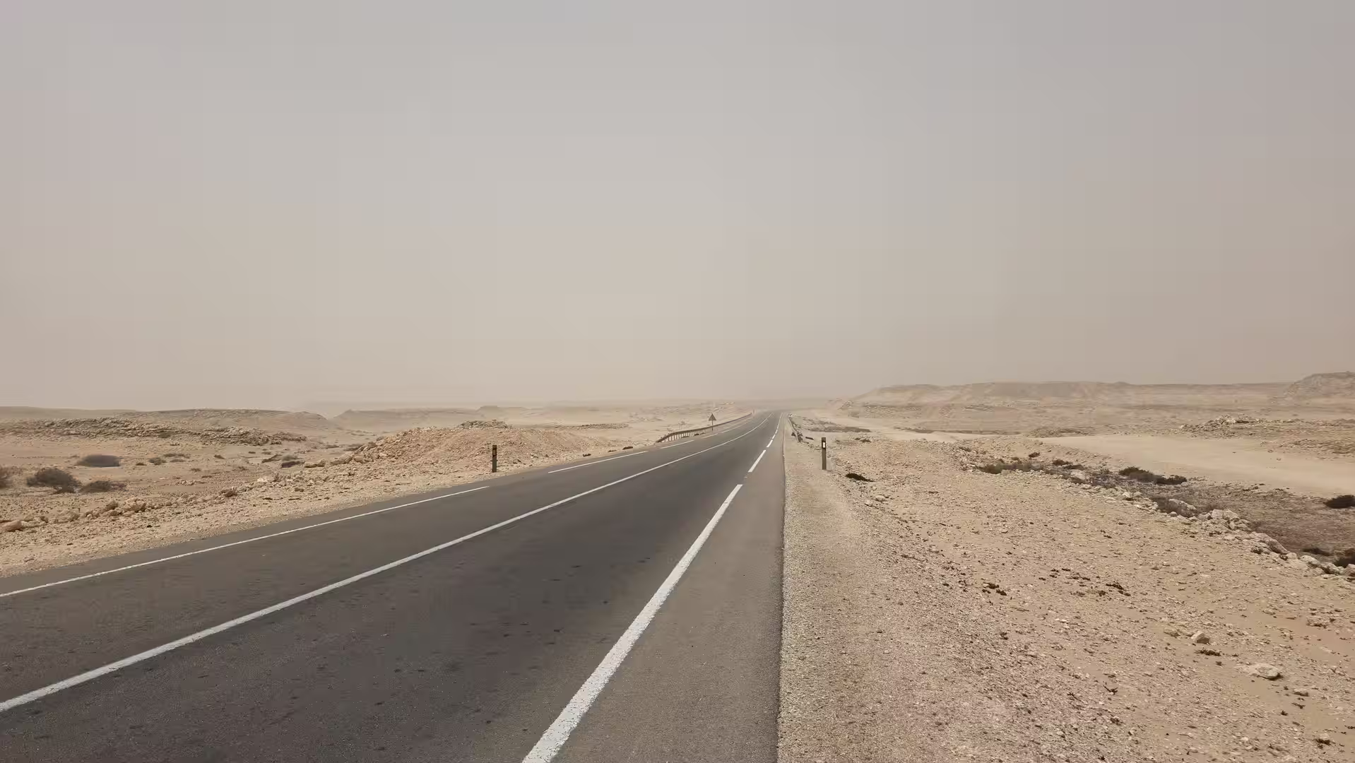 Long paved road crossing a rocky desert plateau under dusty and hazy conditions in Western Sahara.