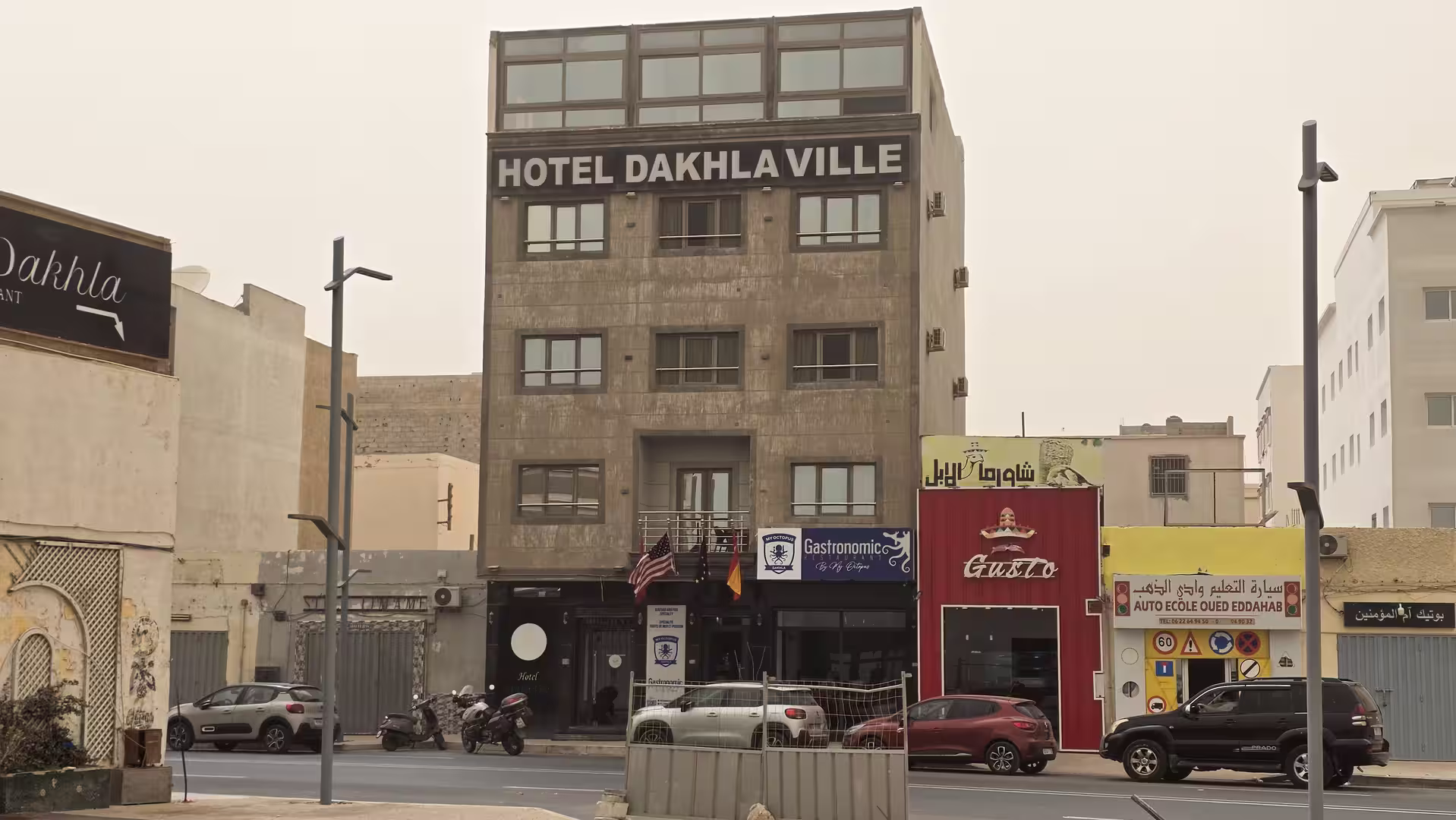 Street view of Hotel Dakhla Ville building in Dakhla with parked vehicles and neighboring storefronts.