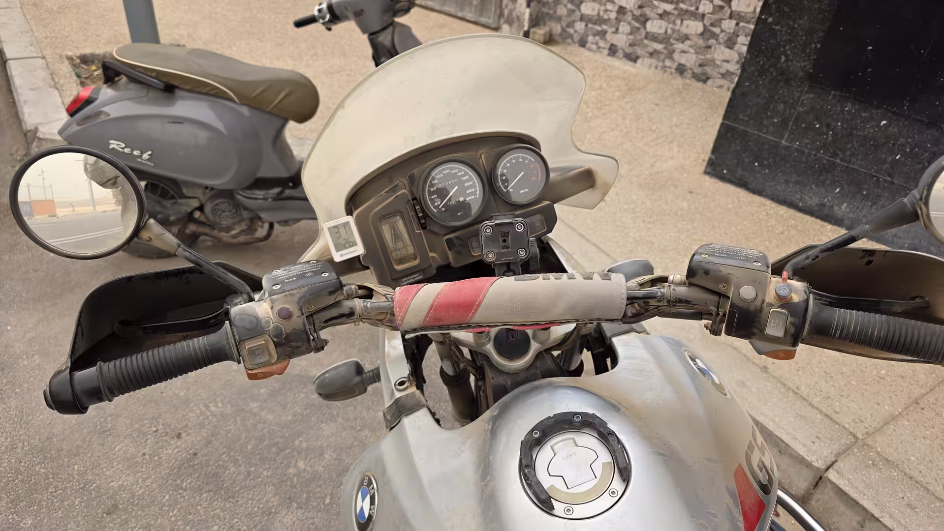 View over the handlebars and instrument cluster of a dusty BMW R 1150 GS parked on a street in Dakhla.