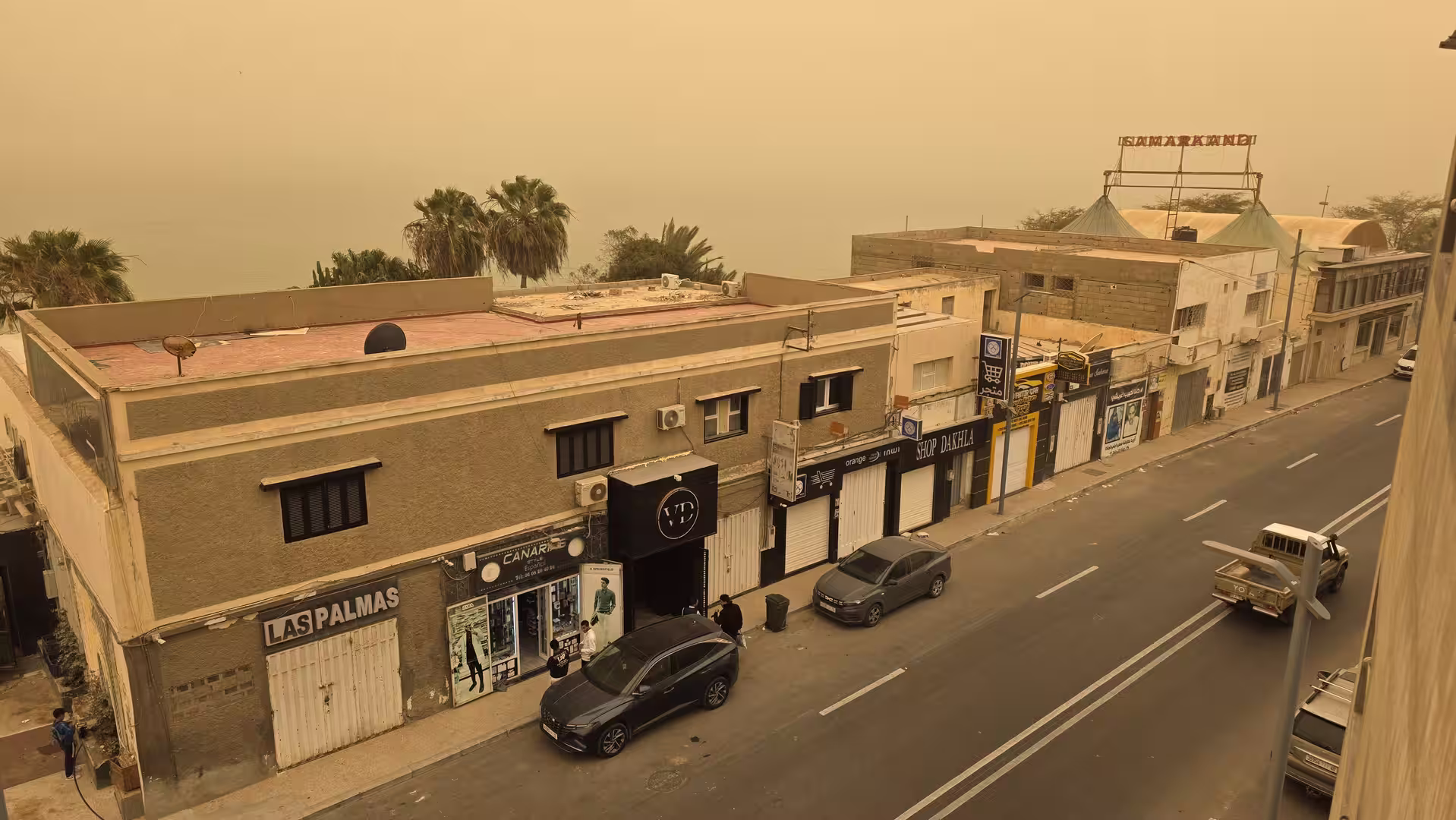 View from an upper floor over a street in Dakhla with shops, parked cars and heavy orange dust reducing visibility toward the lagoon.