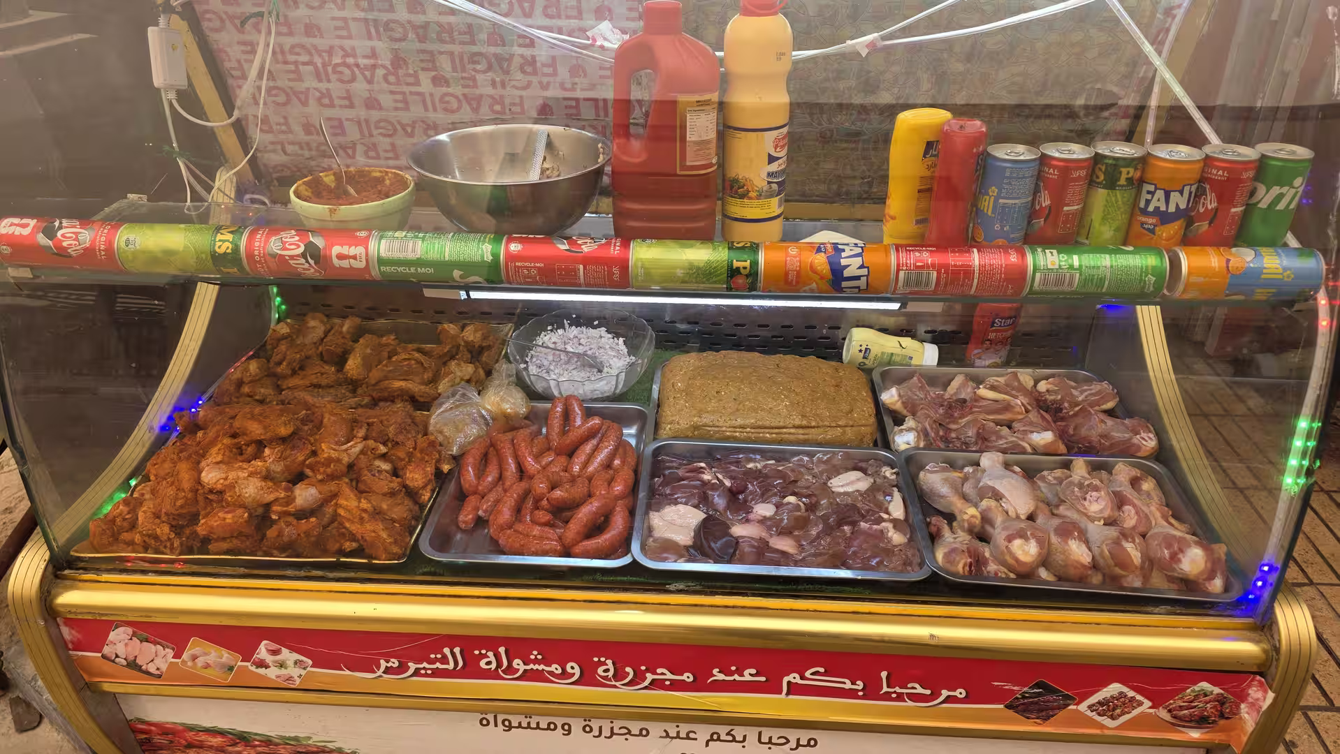 Glass display case in a local eatery showing marinated chicken, sausages and assorted raw meats with condiments and soft drinks above.