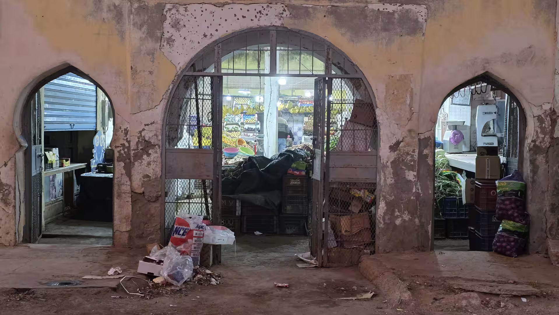 Weathered arched market entrance opening into brightly lit fruit and vegetable stalls in Dakhla at night.