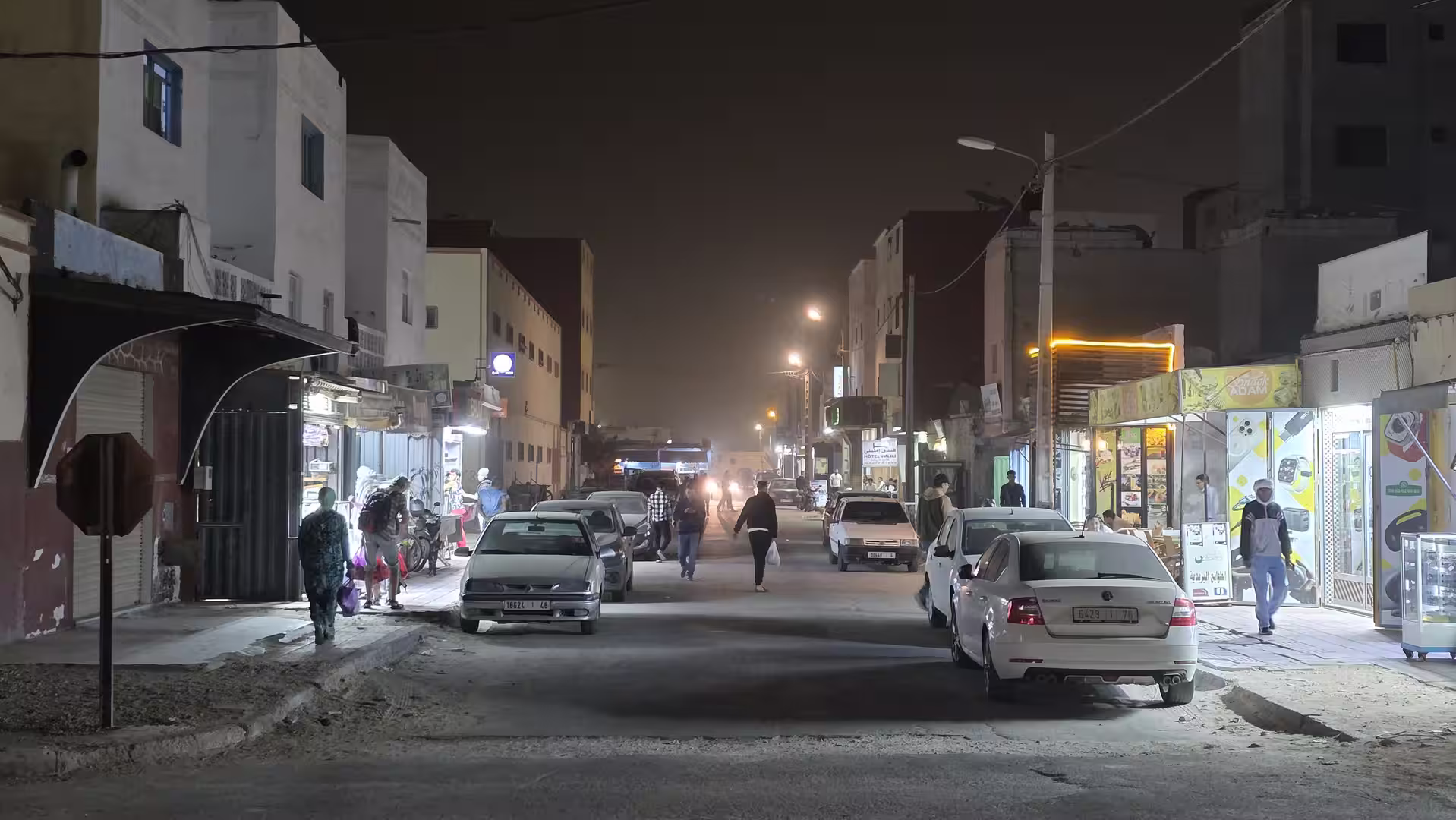 Urban street in Dakhla at night with parked cars, illuminated shops and pedestrians moving through the neighbourhood.