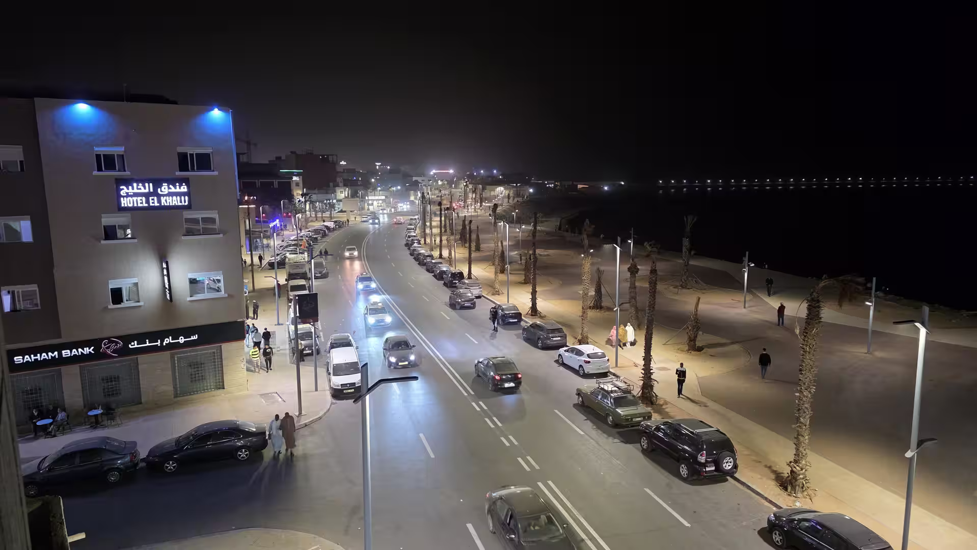 Elevated night view over Dakhla’s illuminated waterfront road with cars, pedestrians and a coastal promenade.
