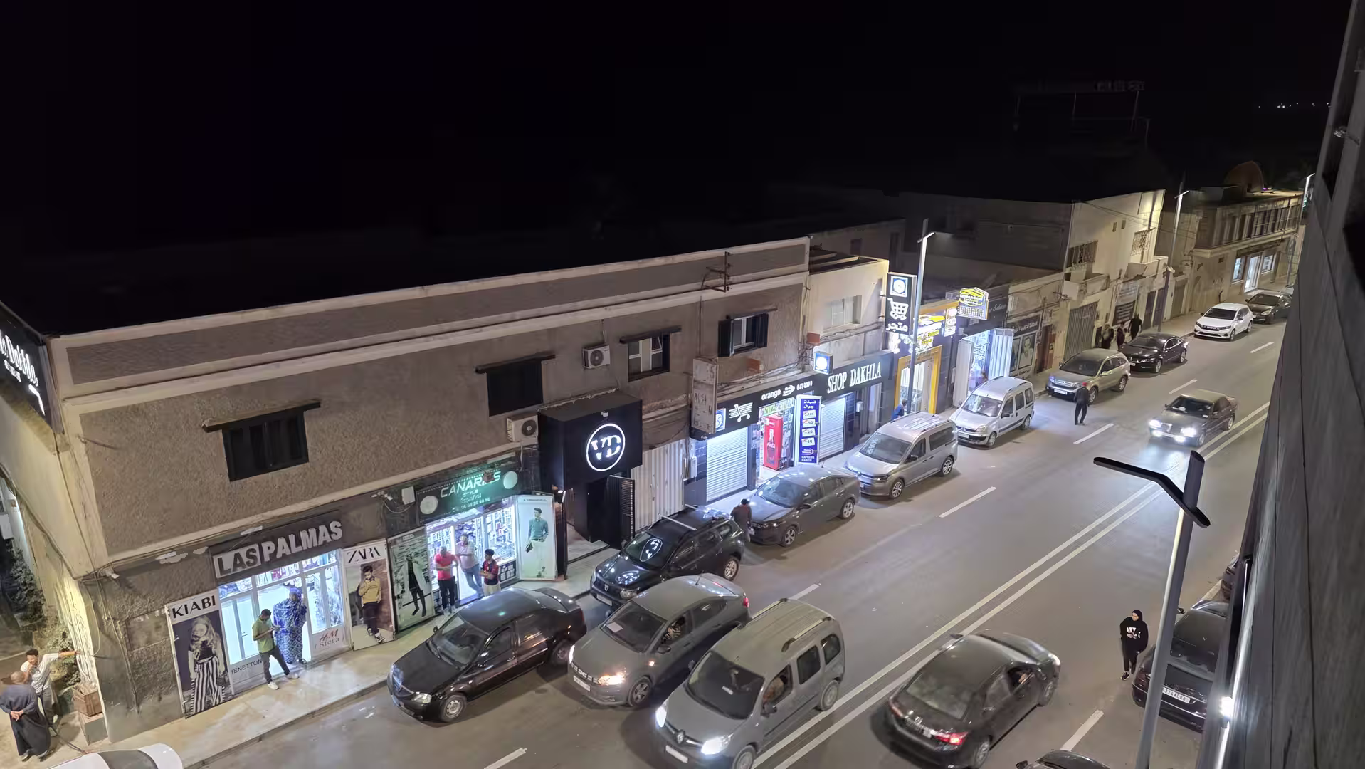 Elevated night view of a commercial street in Dakhla with illuminated shops, parked cars and evening traffic.
