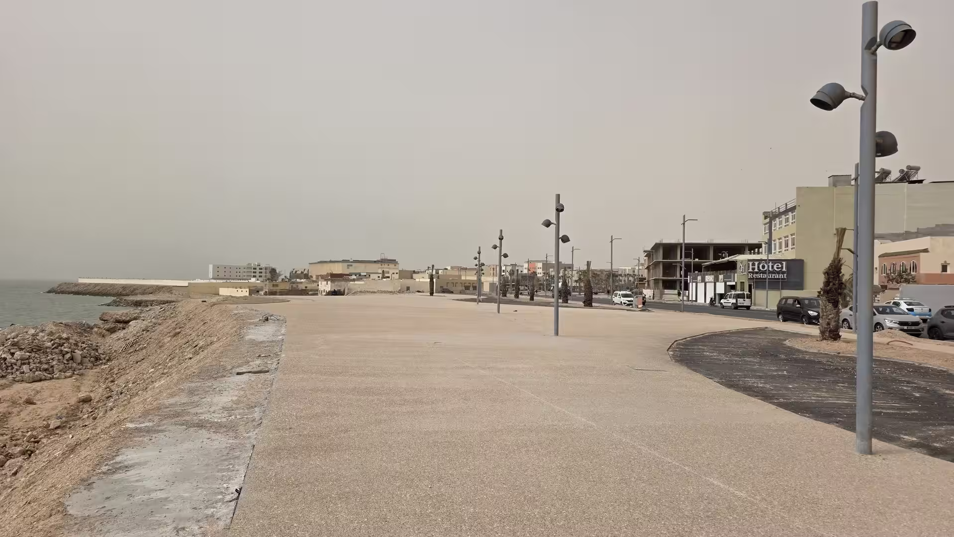 Wide pedestrian promenade along the coast in Dakhla with modern street lighting, a seawall, hotels and new urban development under a dusty sky.