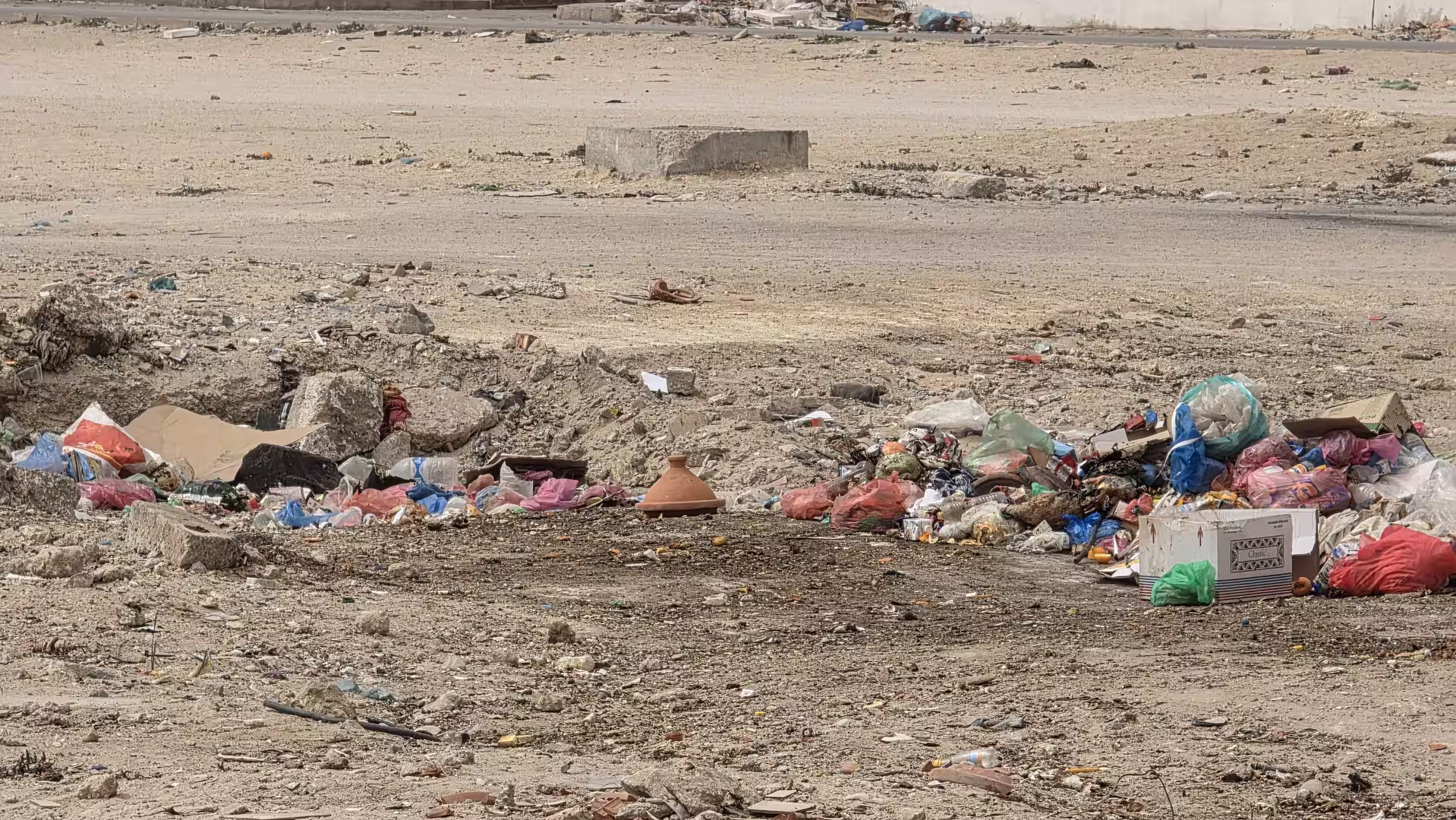 Scattered household waste and plastic bags dumped on barren sandy ground in an undeveloped urban area.