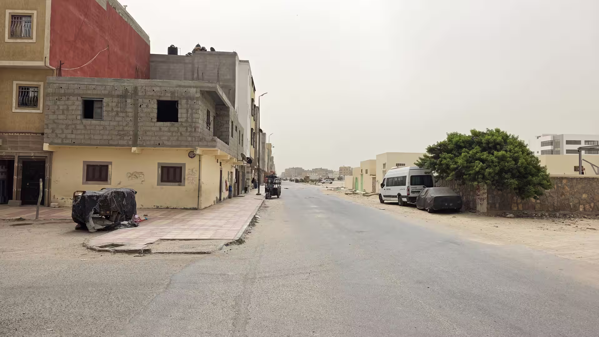 Quiet residential street with unfinished buildings, sandy roadside areas and scattered vegetation under a hazy sky.
