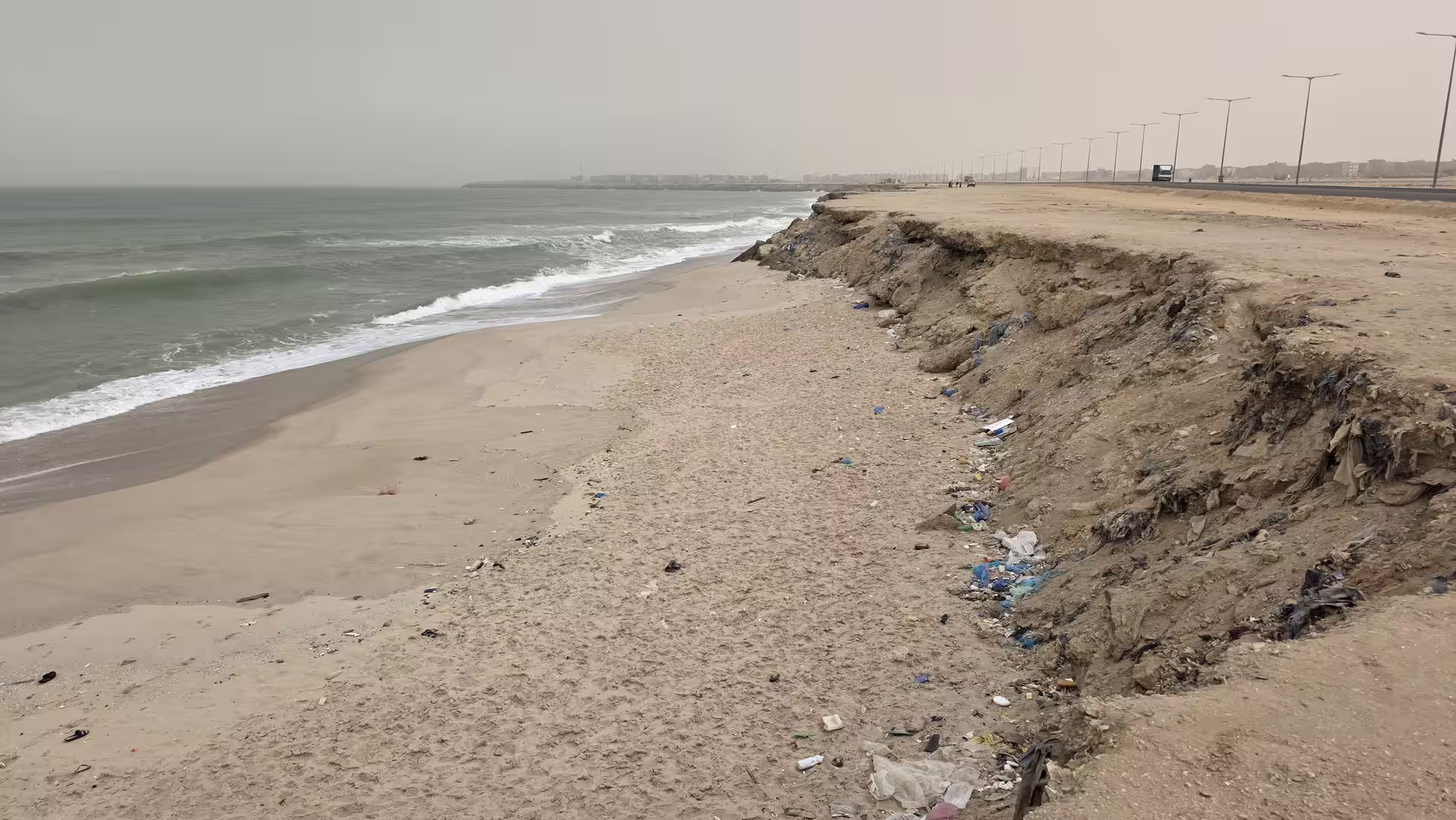 Sandy beach with visible coastal erosion, scattered plastic waste and a road running parallel to the shoreline under hazy conditions.