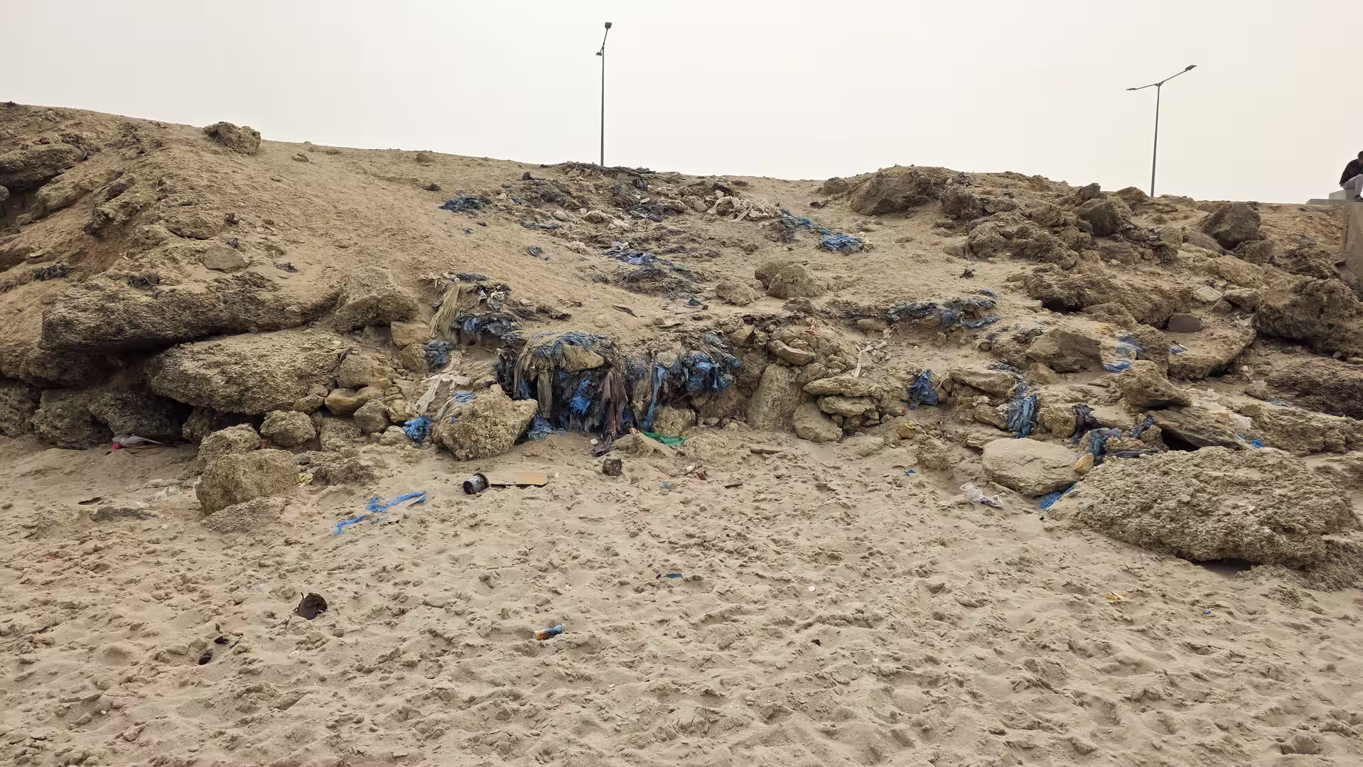 Eroded sandy and rocky coastal edge with exposed layers containing trapped blue plastic waste and debris.