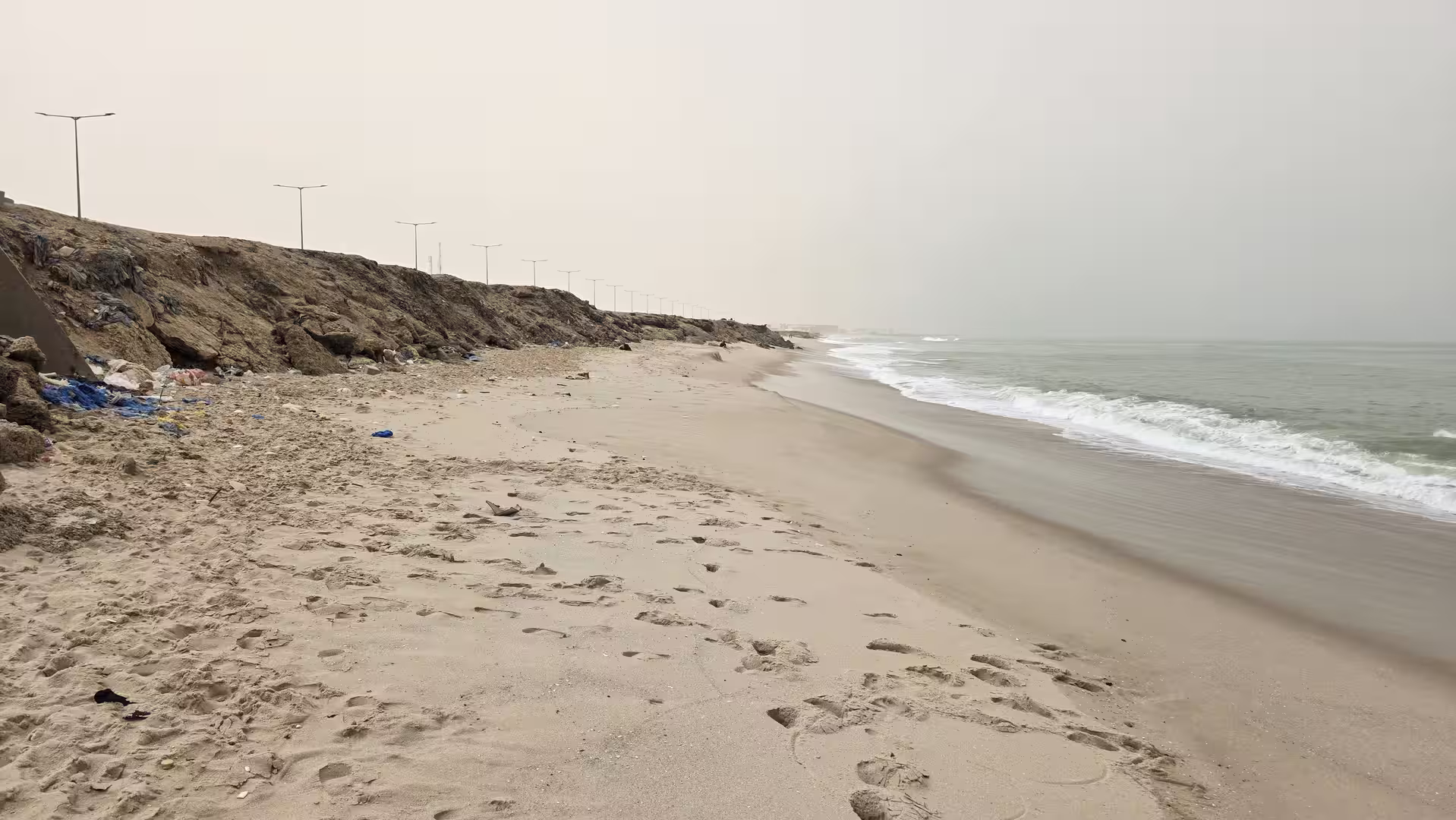 Sandy beach with wave wash, footprints and an eroded coastal embankment running below an urban waterfront road.