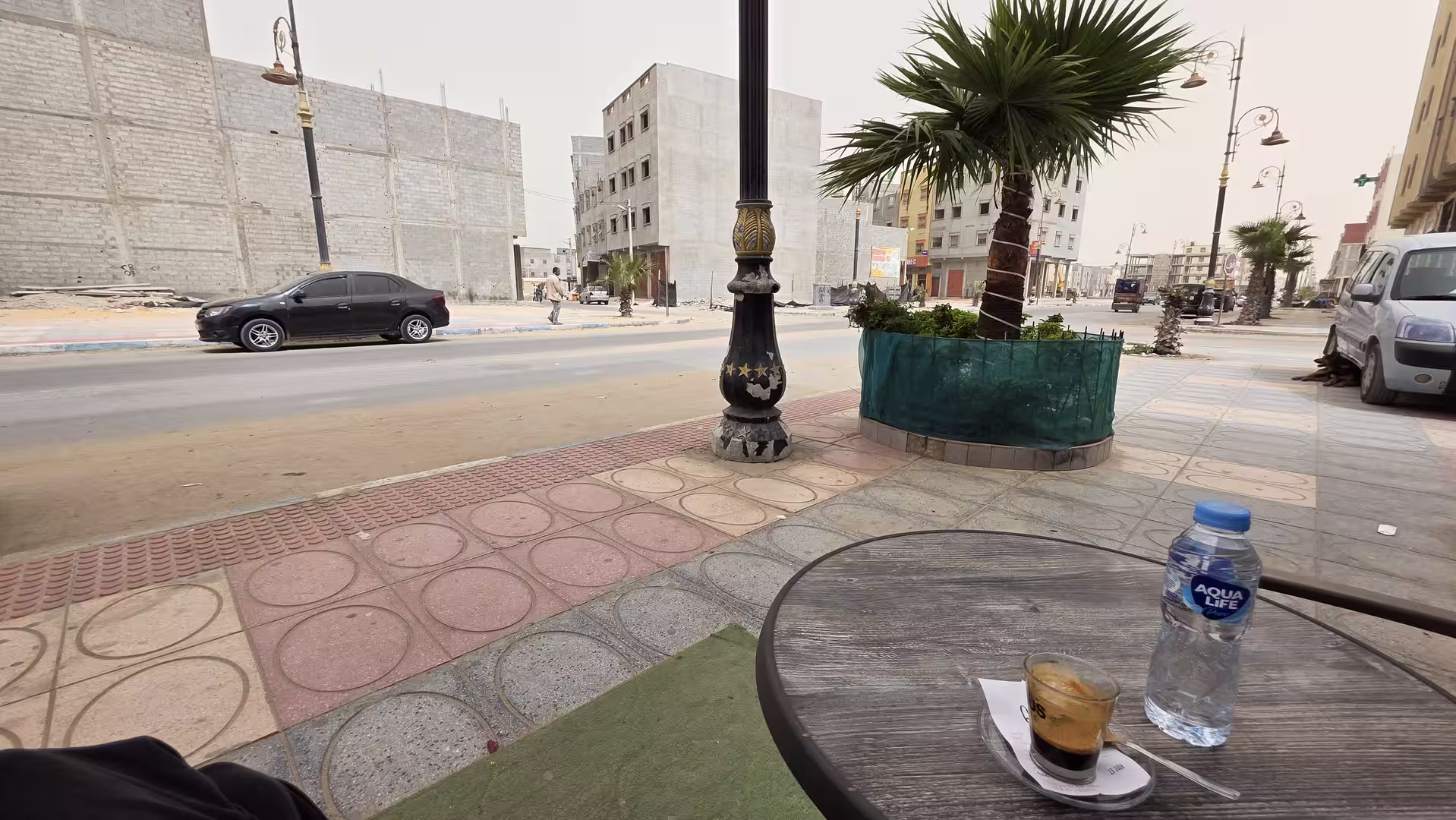 Small espresso and bottled water on an outdoor café table beside a palm-lined street in Dakhla.