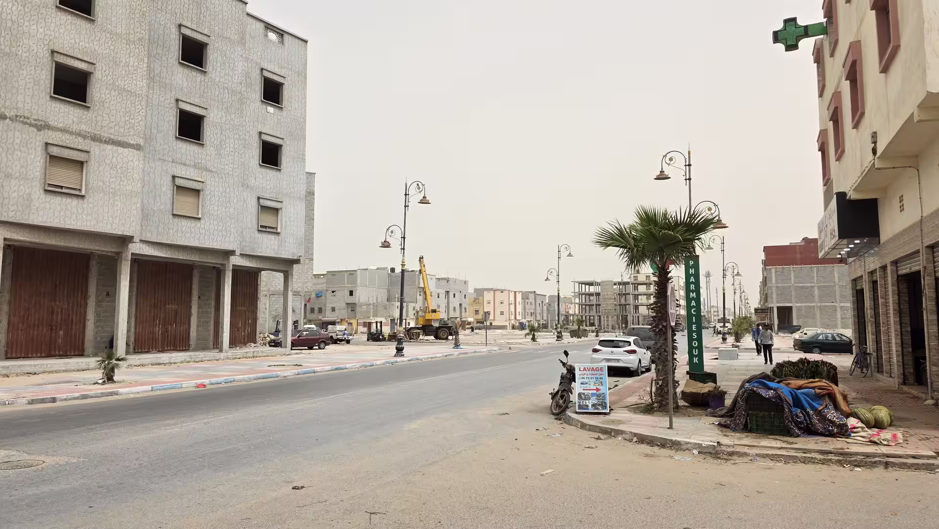 Wide city street in Dakhla with palm trees, new buildings, decorative street lamps and a pharmacy sign.