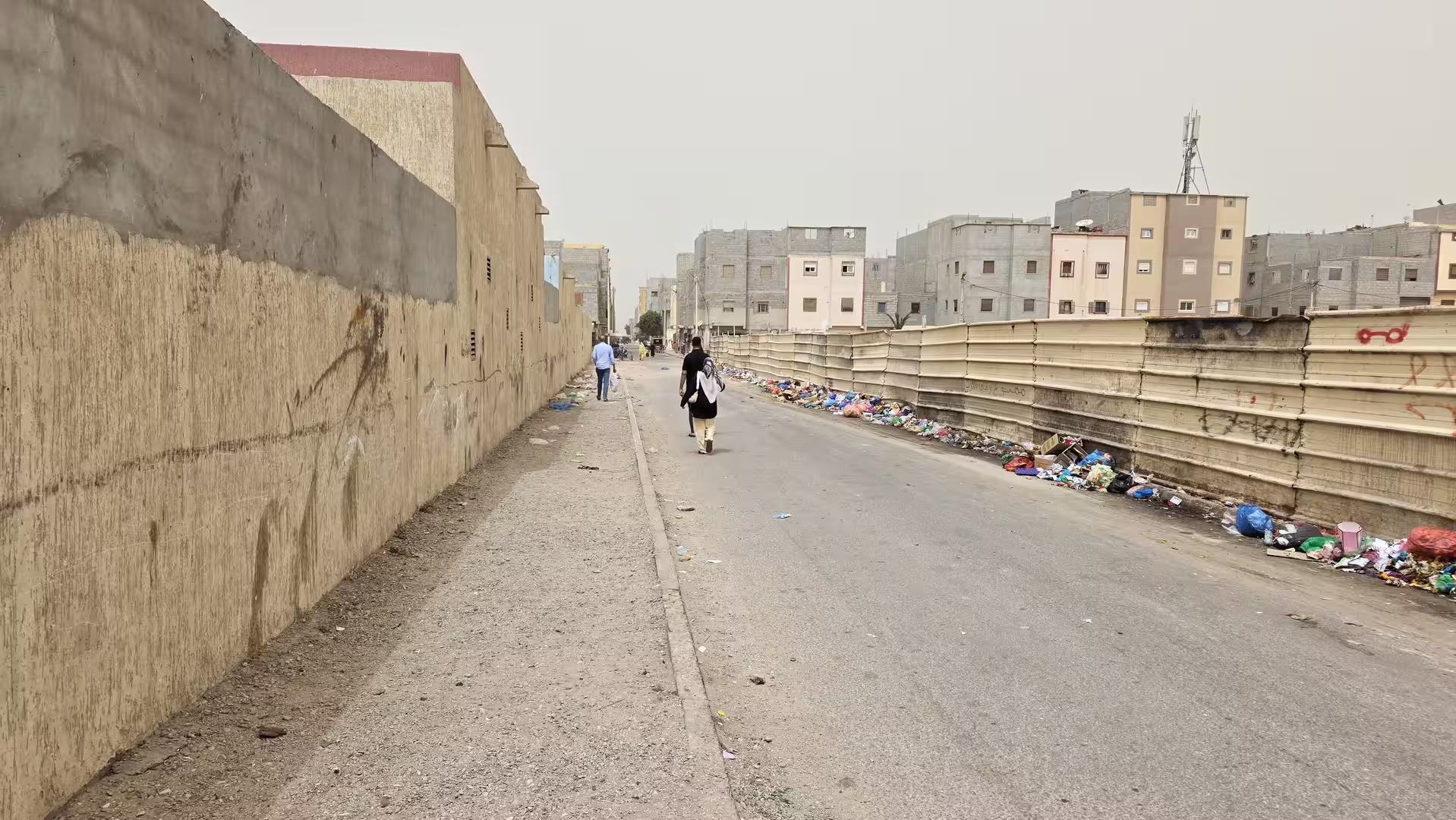 Long narrow street in Dakhla bordered by walls and fencing with scattered waste along one side.