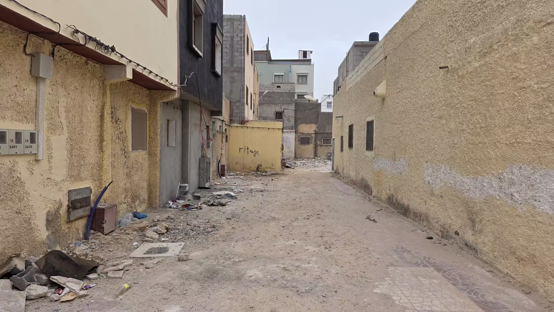 Narrow unpaved alley between residential buildings with scattered construction debris and rough surfaces in Dakhla.