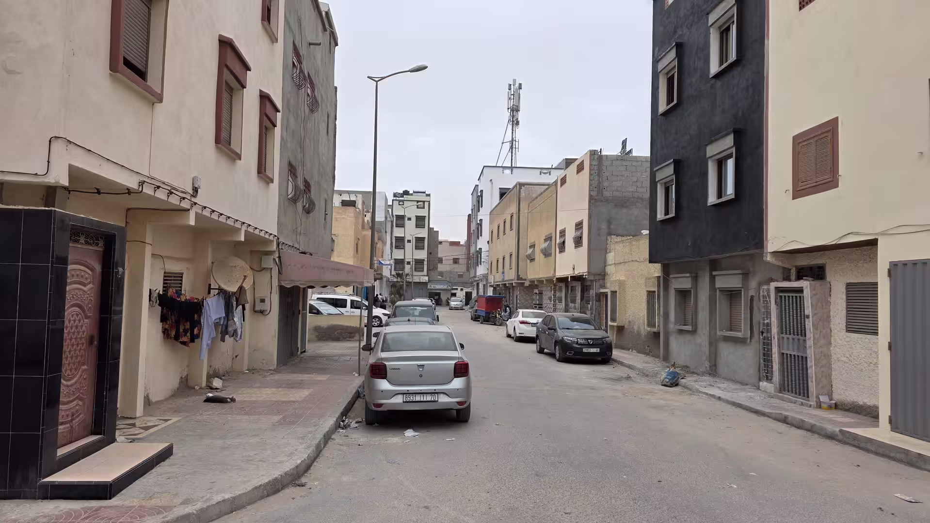 Quiet residential street in Dakhla lined with mid-rise apartment buildings, parked cars and narrow sidewalks.