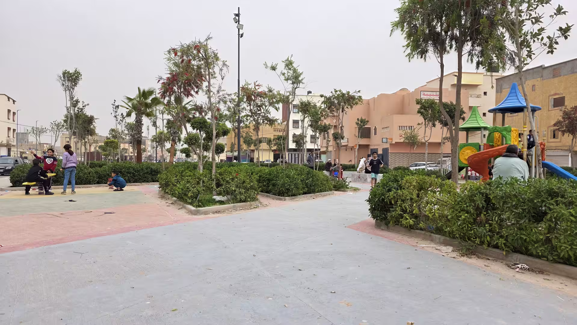 Small urban park in Dakhla with landscaped paths, flowering trees, children playing and a colorful playground.