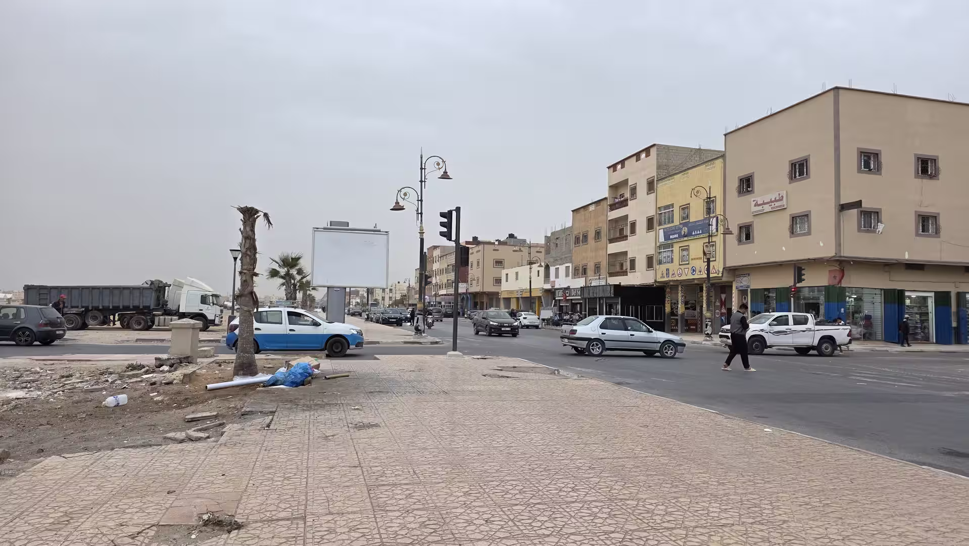 Wide intersection in Dakhla with low-rise buildings, moving traffic, pedestrians and a broad tiled sidewalk.