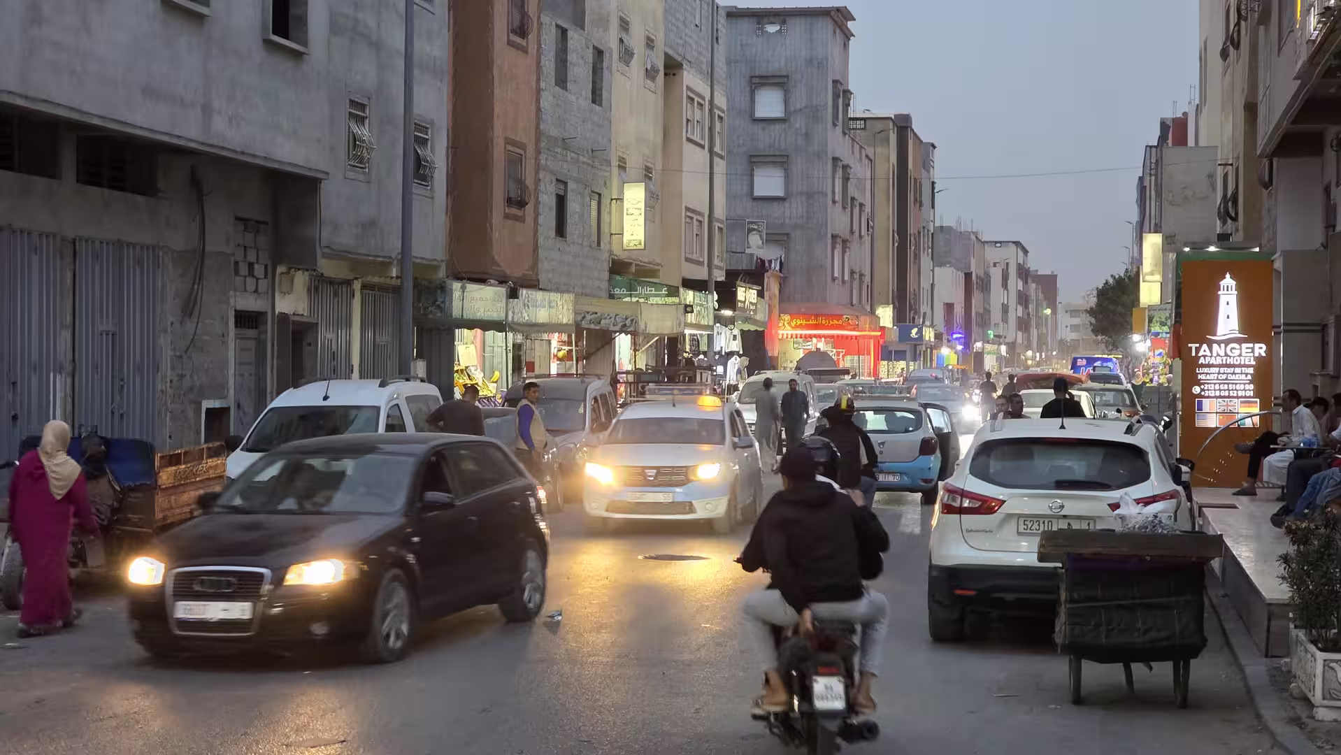 Busy urban street in Dakhla at dusk with cars, motorcycles, illuminated storefronts and pedestrians.