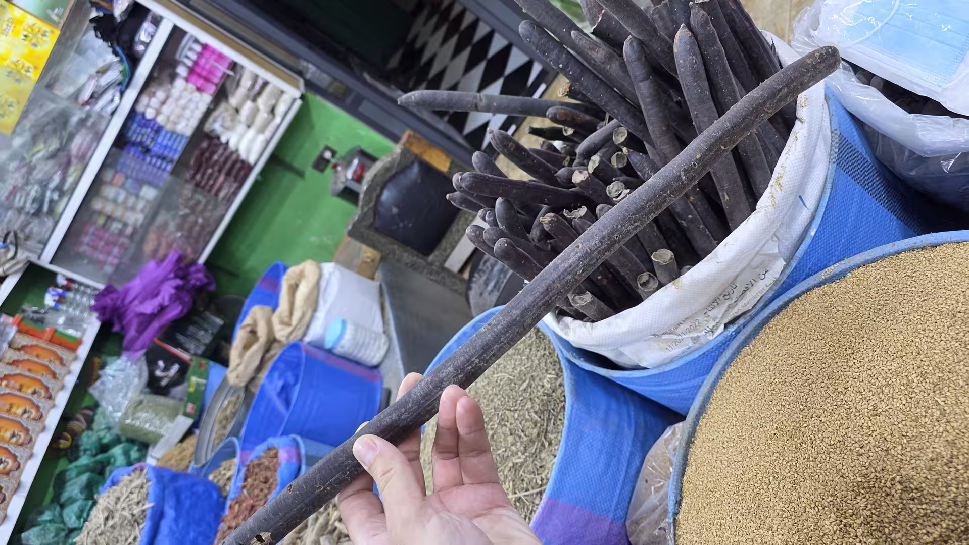 Close-up of long dark dried pods displayed in sacks at a local market stall alongside seeds and spices.