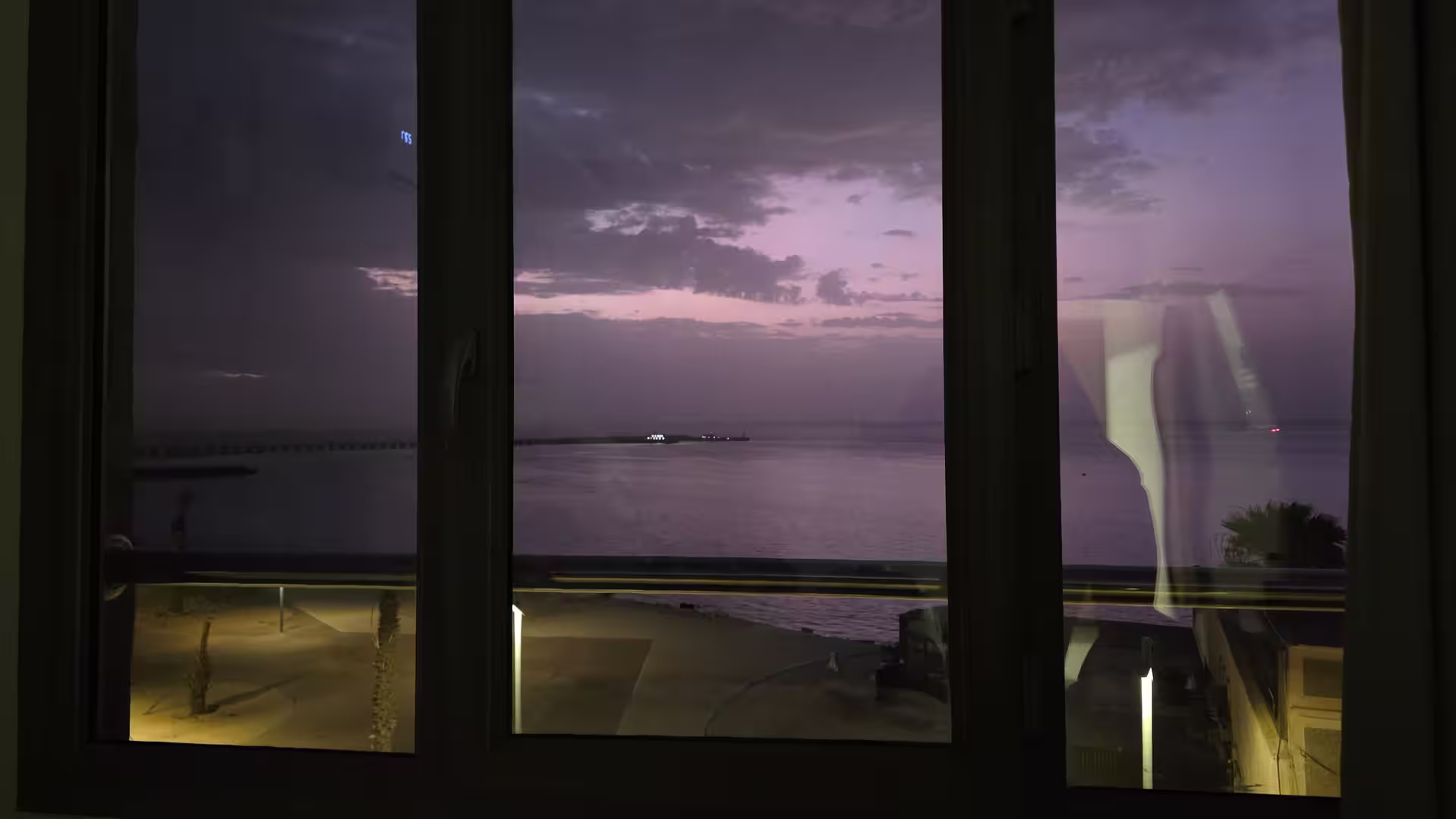 View through a hotel window over Dakhla’s Atlantic waterfront at dusk, showing calm coastal waters, a purple evening sky and softly illuminated promenade below.