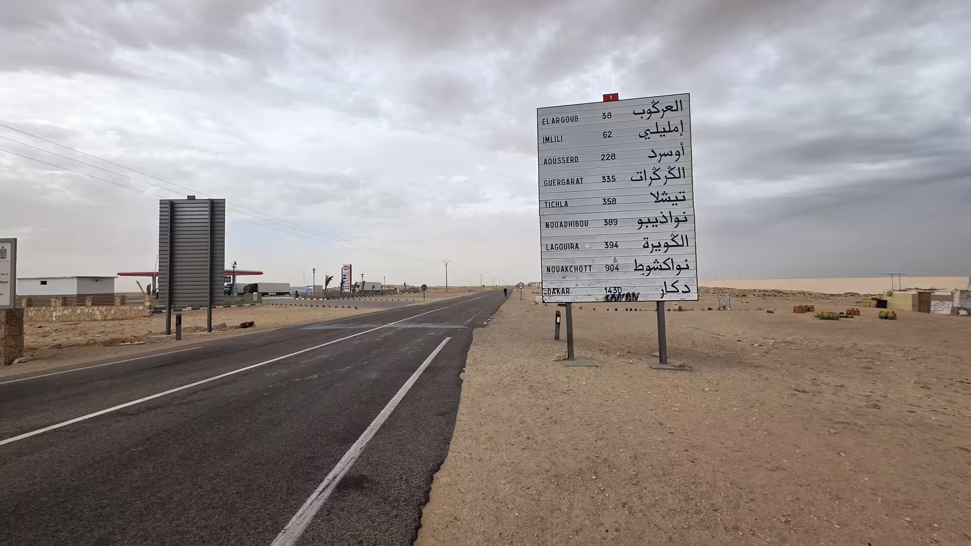 Road sign in the desert south of Dakhla showing distances to destinations in southern Morocco and Mauritania under a dramatic cloudy sky.