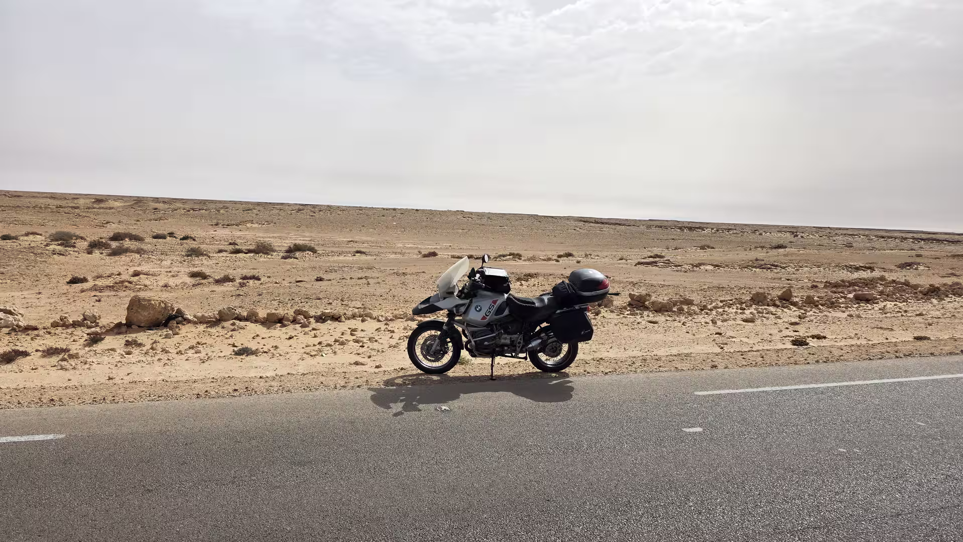BMW GS motorcycle parked beside a remote desert road during a northbound journey through the Sahara landscape near Dakhla under a bright sky.