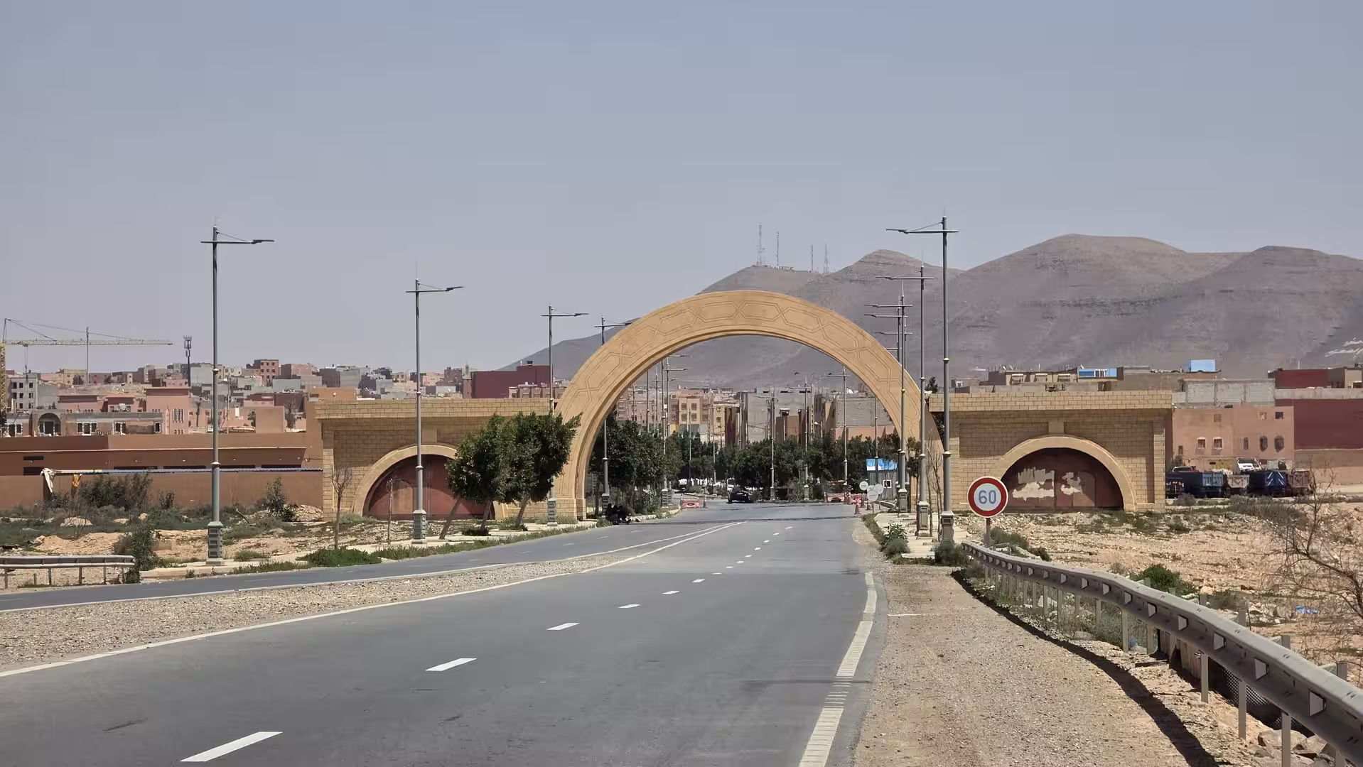 A wide road leads through a large stone gateway at the entrance of a desert city, with low-rise buildings and rocky hills rising in the background under a clear sky.