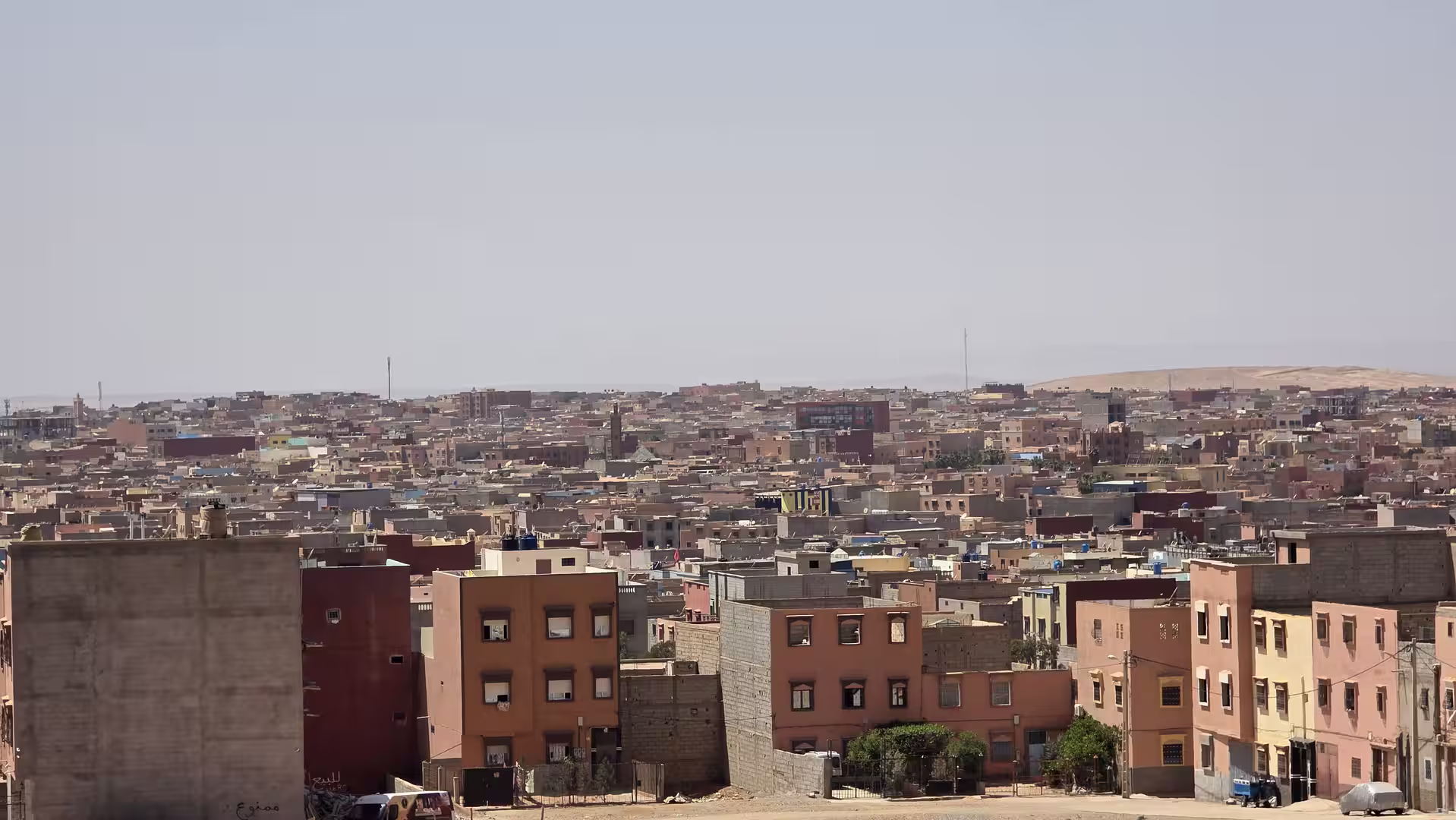 Panoramic view over a desert city with densely packed low-rise buildings extending toward sandy hills under a clear sky.