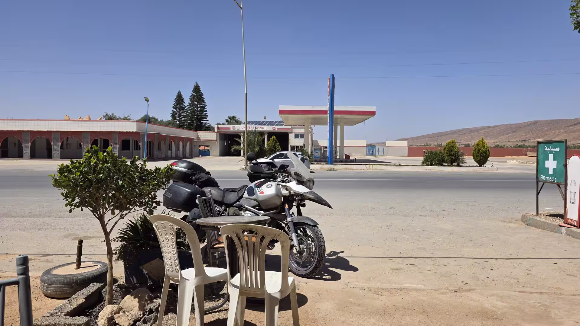 A touring motorcycle parked beside outdoor café chairs on a roadside stop in southern Morocco, with a fuel station and dry hills in the background.