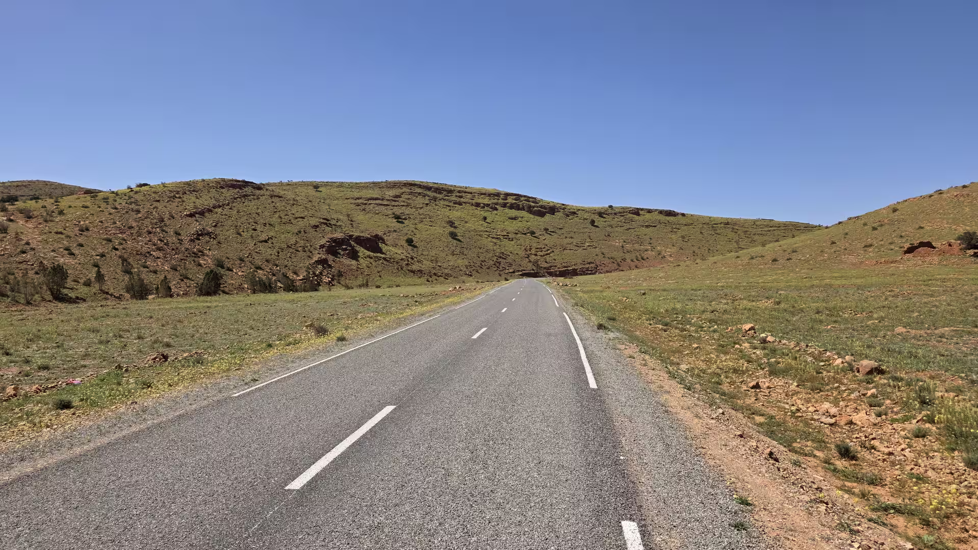 A quiet road winds through green hills and open countryside beneath a clear blue sky in southern Morocco.