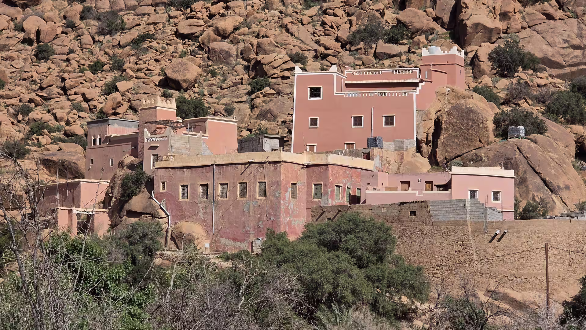 Pink and earth-toned houses are built among large rock formations in a mountain village in Morocco, surrounded by sparse vegetation.