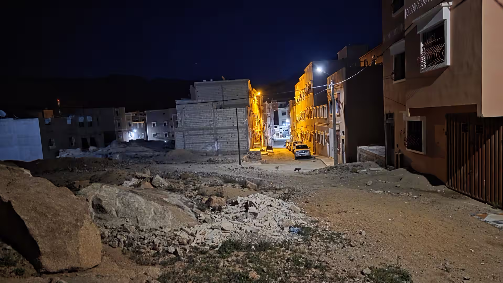 A quiet street in Tafraoute at night, where warm streetlights illuminate residential buildings set among rocky terrain and the surrounding mountains.