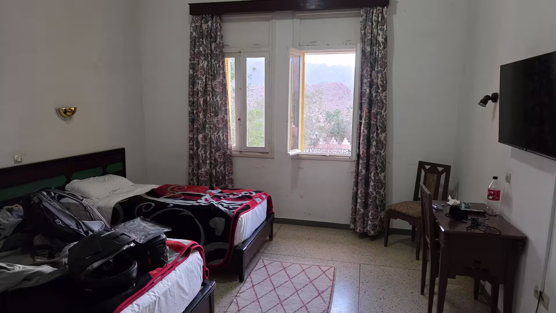 Simple hotel room with twin beds, a desk, TV, and an open window overlooking the mountains.