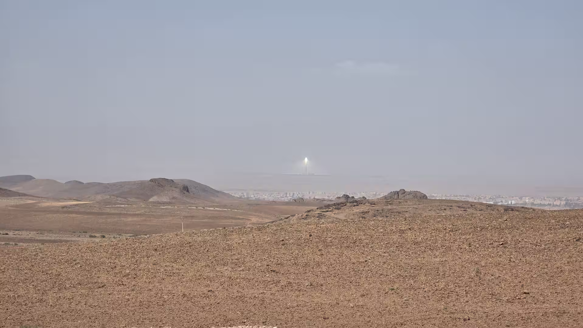 A dry rocky plateau in the Atlas region with low hills and a bright concentrated reflection visible far on the horizon.