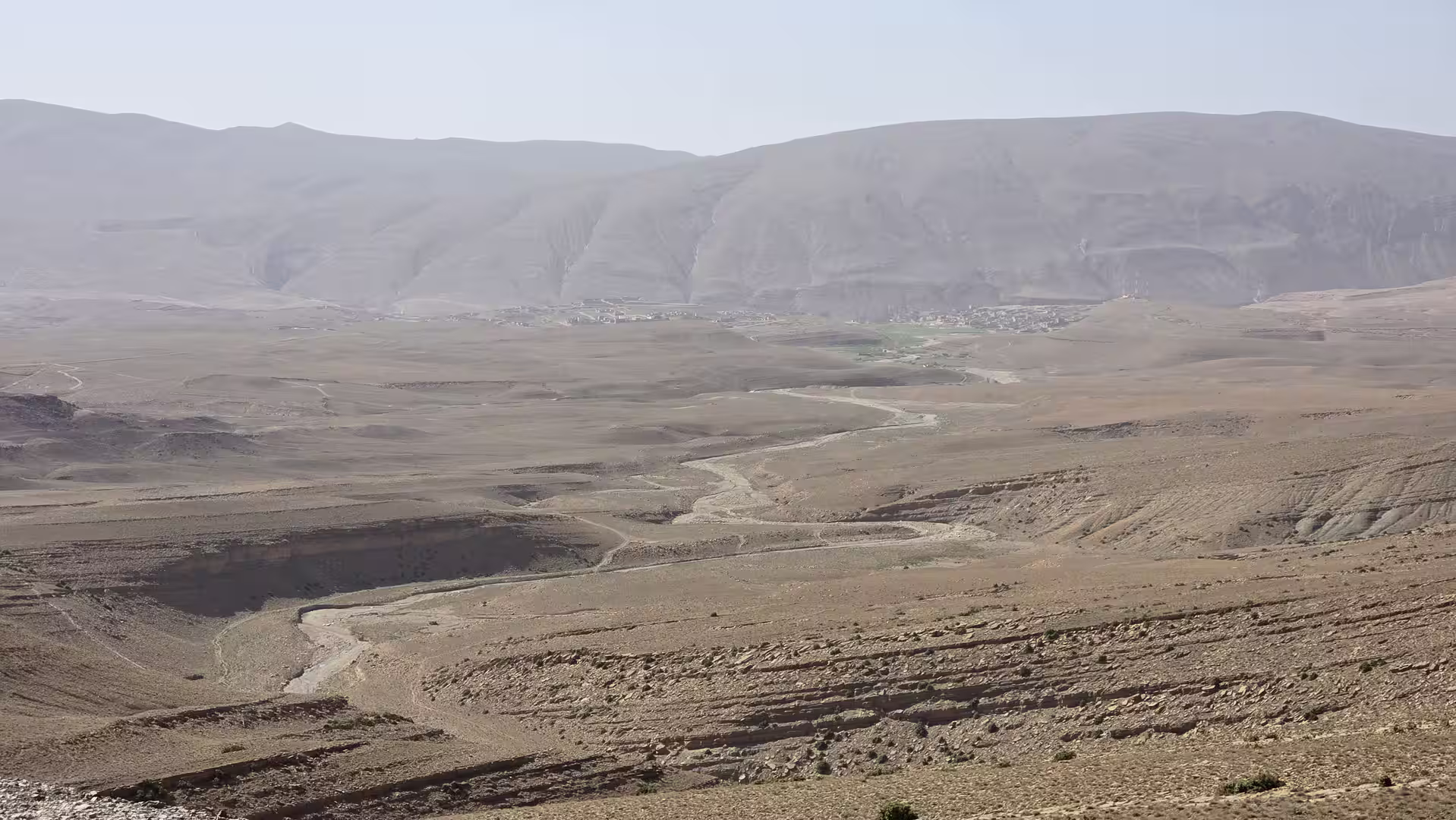 Wide arid mountain basin with a winding riverbed leading toward a distant settlement at the base of broad Atlas mountain slopes.