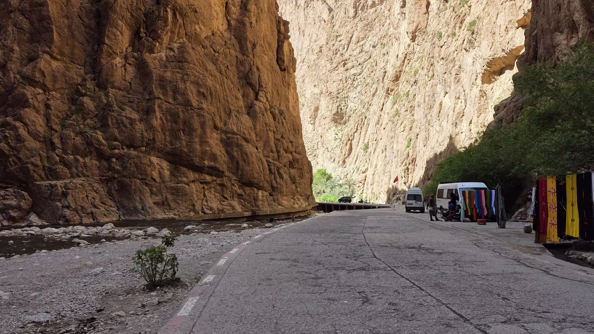 Road running through a narrow canyon with towering rock walls, a flowing stream, roadside market stalls, and parked vehicles.